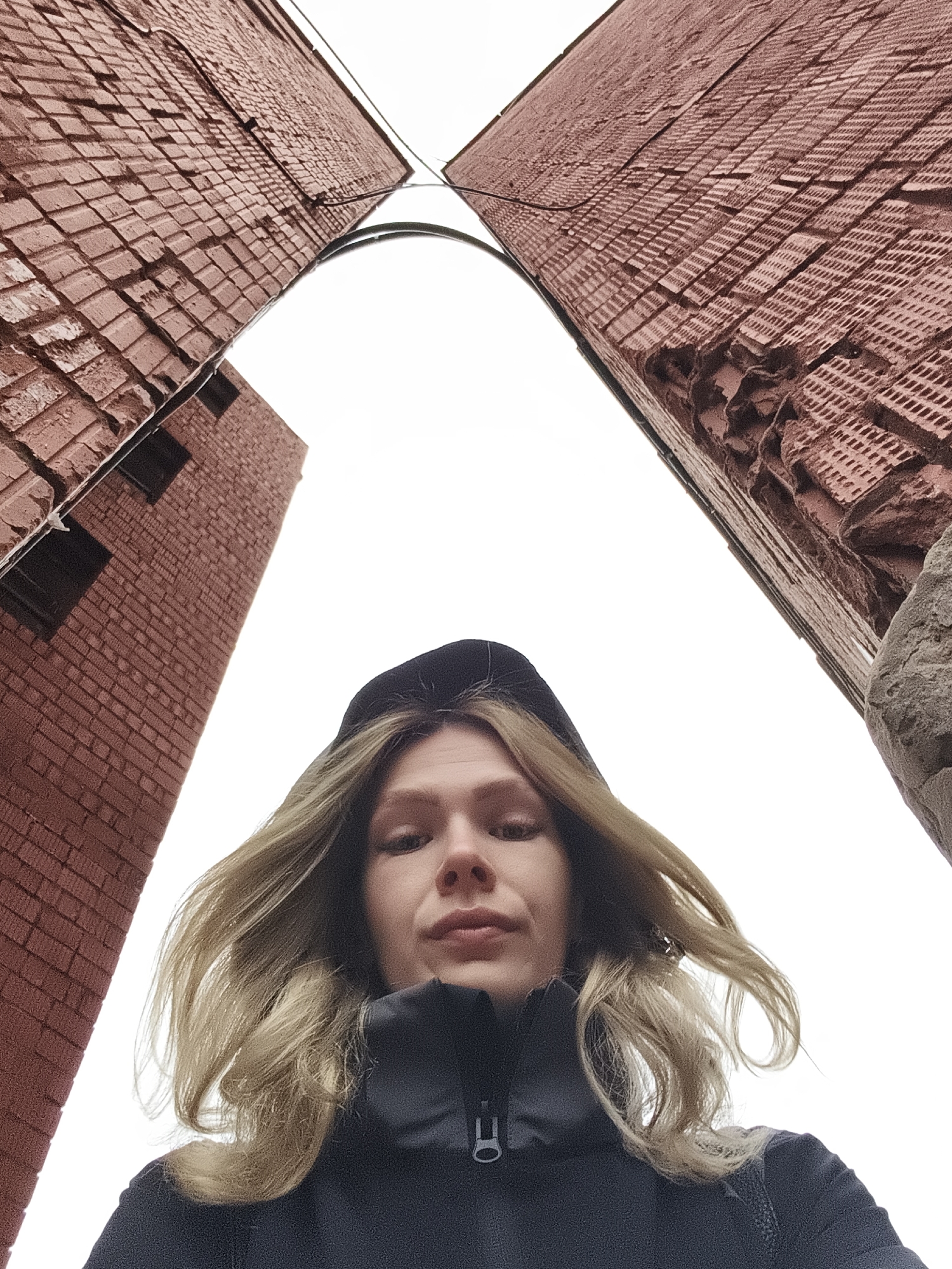 A low-angle photograph shows a person, presumed female, in the foreground, framed by two brick buildings. The person is wearing a dark hooded jacket. Their blonde hair is partially windblown. The buildings on either side are brick, appearing to be a reddish-brown color. The sky is overcast and white. A black cable runs between the tops of the buildings. The architectural style of the brickwork is visible, showing detail in the construction. The image is taken in the city of Izhevsk, Russia.