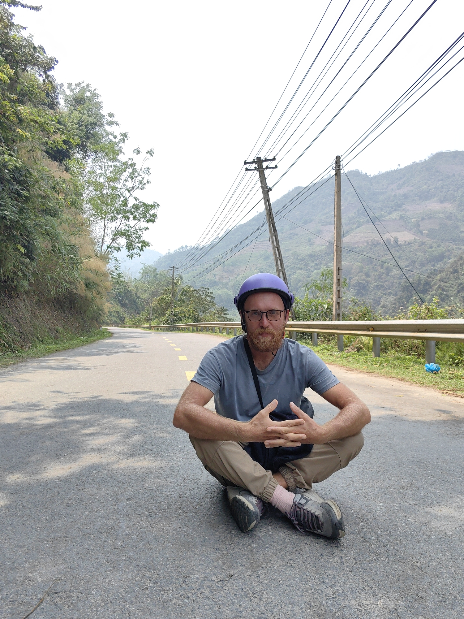 A man wearing a purple helmet, glasses, a gray T-shirt, and khaki pants is sitting cross-legged on a gray asphalt road. His hands are clasped in front of him. He is wearing sneakers and pink socks. Behind him is a metal guardrail, green vegetation, and a hillside with a mountain in the distance. Electrical poles and wires are visible overhead. The road curves in the distance. The scene is lit by sunlight, casting shadows on the road. The location is a road in what appears to be a mountainous area. Based on the prompt the location is in the city of Bảo Lạc, Vietnam.