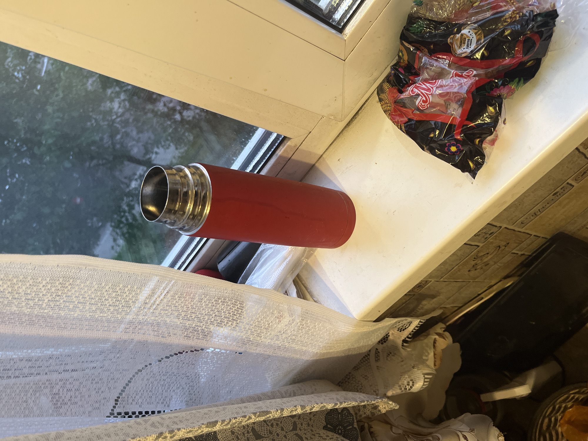 The image depicts a close-up view of a windowsill in Minsk, Belarus. On the windowsill, a bright red thermos is positioned to the left of a white window frame. To the right of the thermos, a crinkled plastic bag with a black background and colorful floral patterns lies on the windowsill. The bag appears to contain food items, with a label visible that includes the text "СМЕХОТА 2022" and "ЖИВОТА". A white sheer curtain hangs on the left side of the frame, partially obscuring the view of the outside, which shows trees and foliage. The overall scene suggests an indoor setting, likely a residential or office space, with a window overlooking an outdoor environment. The lighting is somewhat dim, indicating it might be overcast or evening.