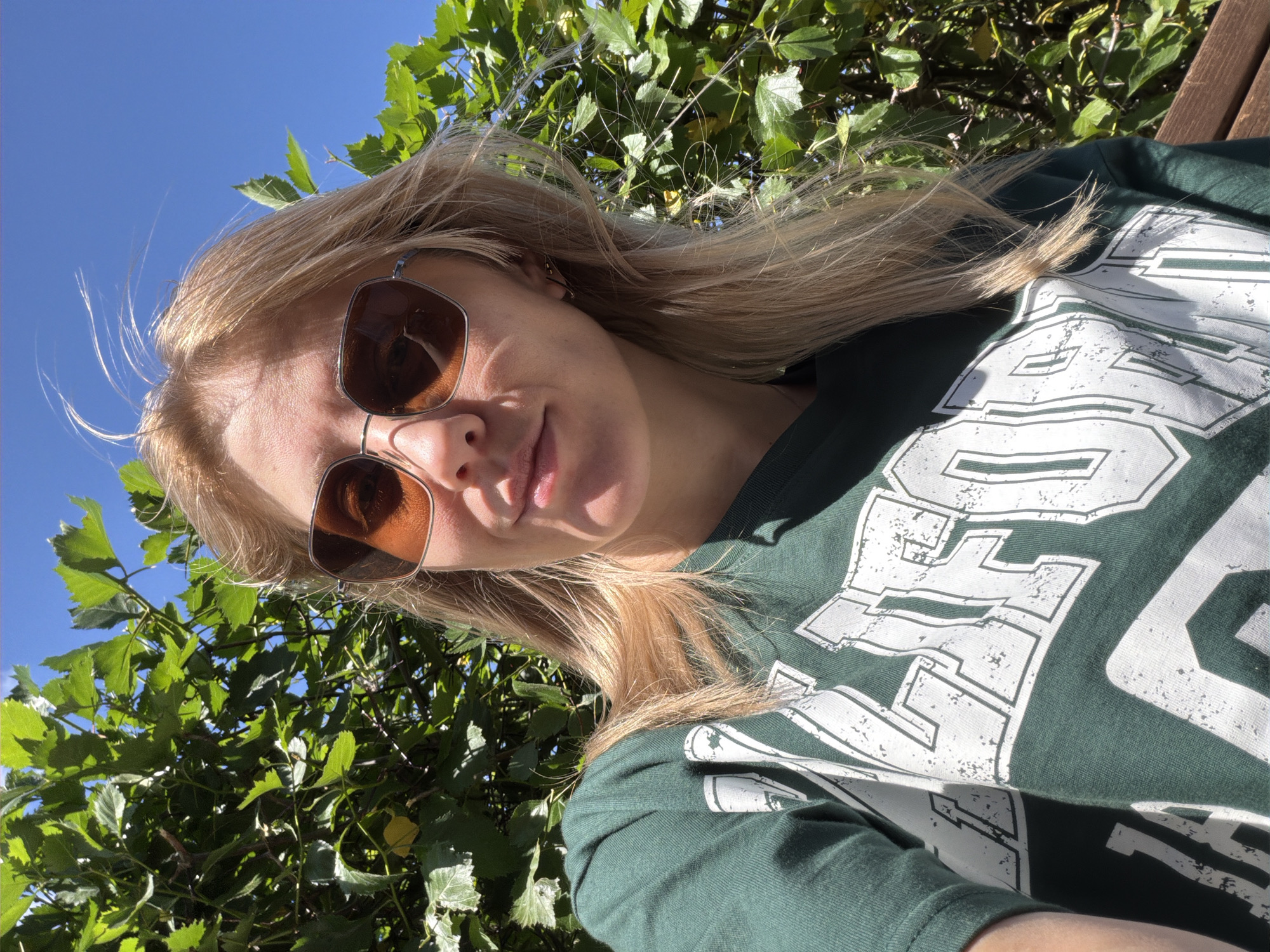 A woman with blonde hair and sunglasses is taking a selfie outdoors. She is wearing a dark green t-shirt with white lettering on it. The t-shirt has a distressed, vintage look. She is sitting on a wooden bench, and behind her is a lush green bush or tree. The sky is bright blue, indicating it is daytime and the weather is sunny. The image appears to be taken during the day, given the strong sunlight and blue sky. The woman's face is partially obscured by sunglasses, but she appears to be smiling faintly. The overall mood is relaxed and casual. There is no visible text that can be read clearly. The image is oriented sideways.