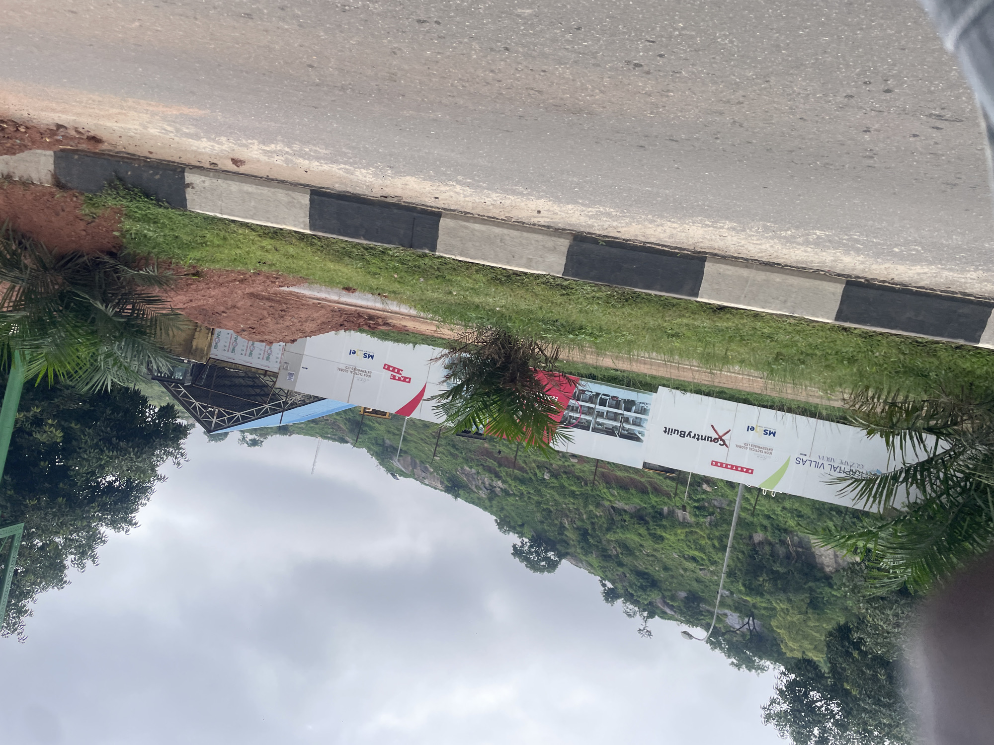 A construction site is visible in Abuja, Nigeria, with a large billboard displaying "CountryBuilt" and other branding. The site is situated at the base of a lush, green, and rocky hillside under a cloudy sky. In the foreground, a paved road with a black and white bordered curb runs parallel to the construction zone. Scattered red soil mounds and green grass cover the immediate area behind the curb. A white temporary structure with a blue roof and exposed beams is also present on the site. Palm trees and other greenery are interspersed throughout the scene.