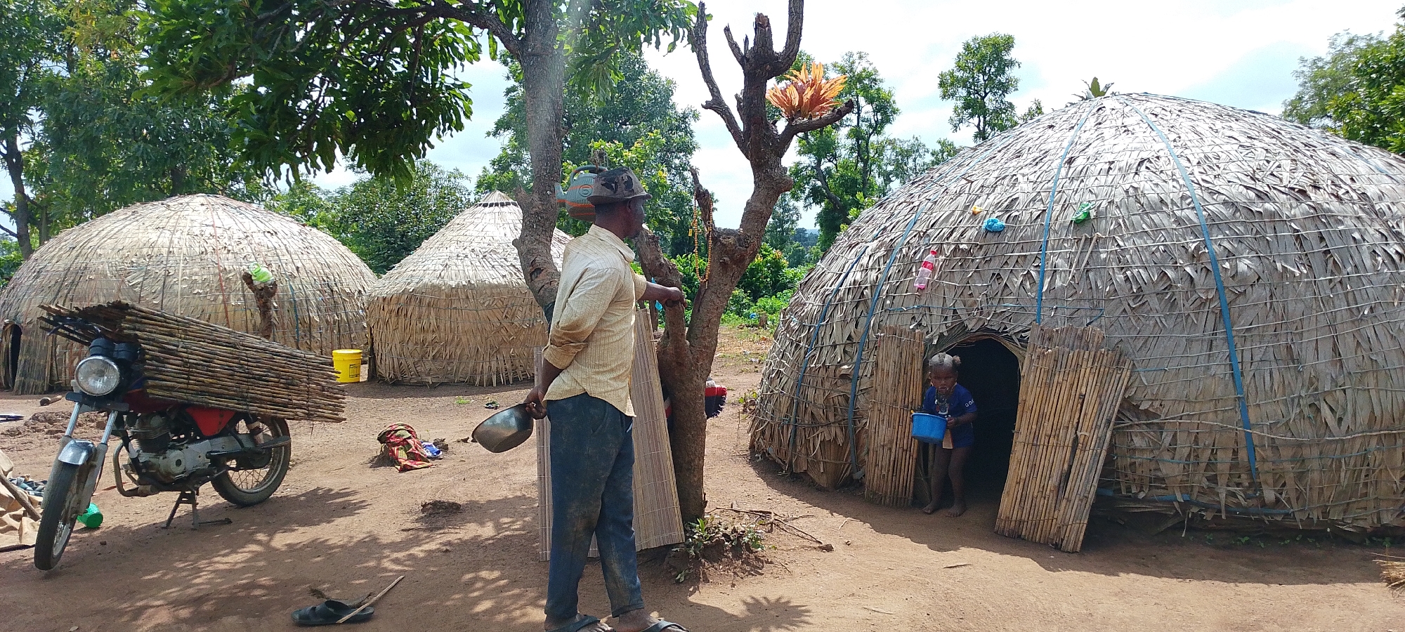 In Kamantan, Nigeria, a rural scene unfolds. Two thatched-roof, conical huts, constructed from dried vegetation, dominate the landscape. A motorcycle, laden with a bundle of bamboo poles, is parked in the foreground. A man, dressed in a light-colored striped shirt and dark pants, stands by a tree, holding a metal bowl. A young child emerges from the doorway of the larger hut, holding a blue cup. The ground is composed of dry, reddish soil, with scattered debris. The environment is lush with green trees, suggesting a tropical or sub-tropical climate. The scene captures a moment of daily life in a remote Nigerian village.
