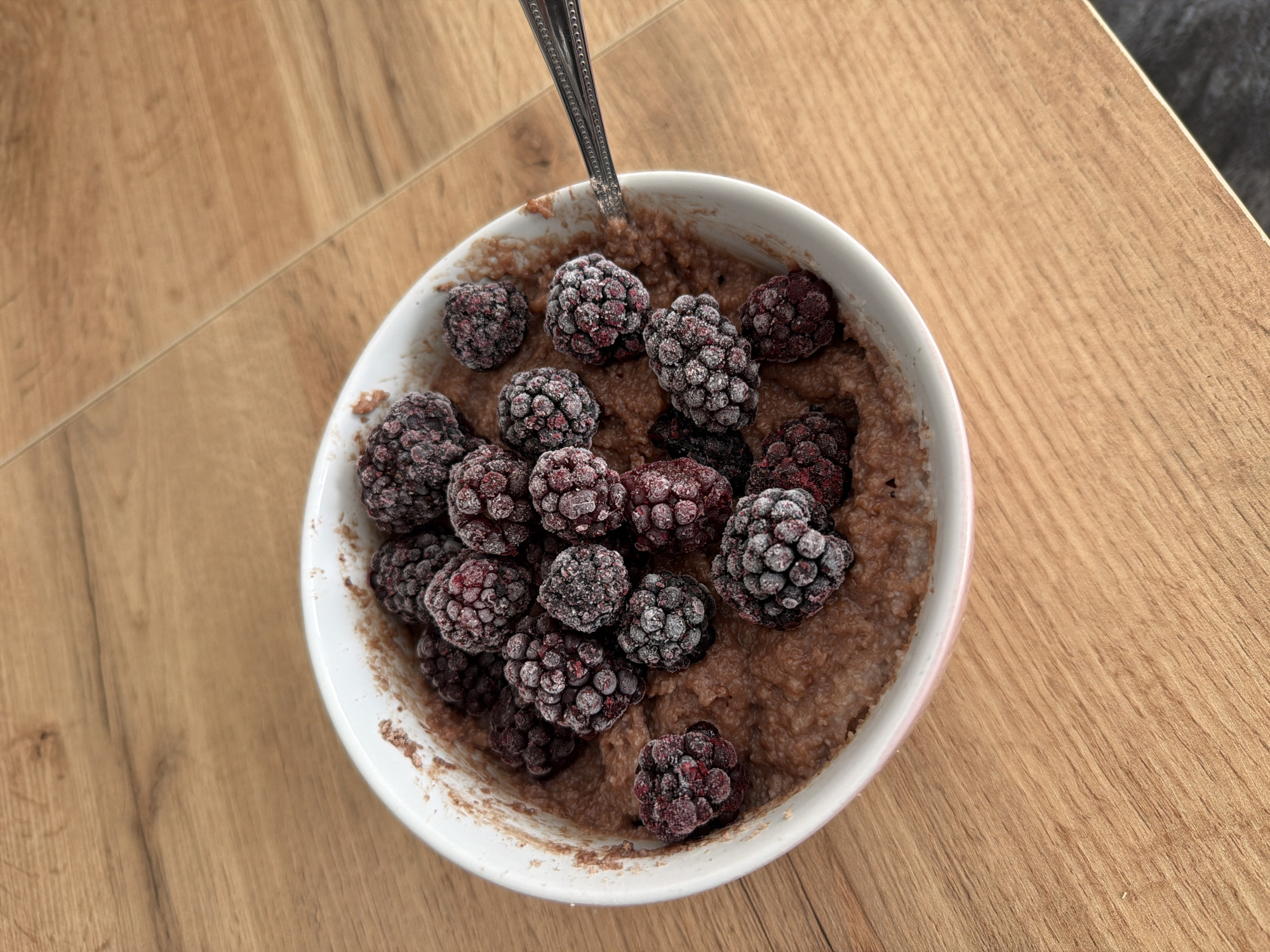 A bowl of oatmeal with blackberries sits on a wooden table. A spoon is in the bowl, ready to be used. The scene is set outdoors, with natural light illuminating the food. There are no people or other objects in the frame. The location is Plovdiv, Bulgaria.
