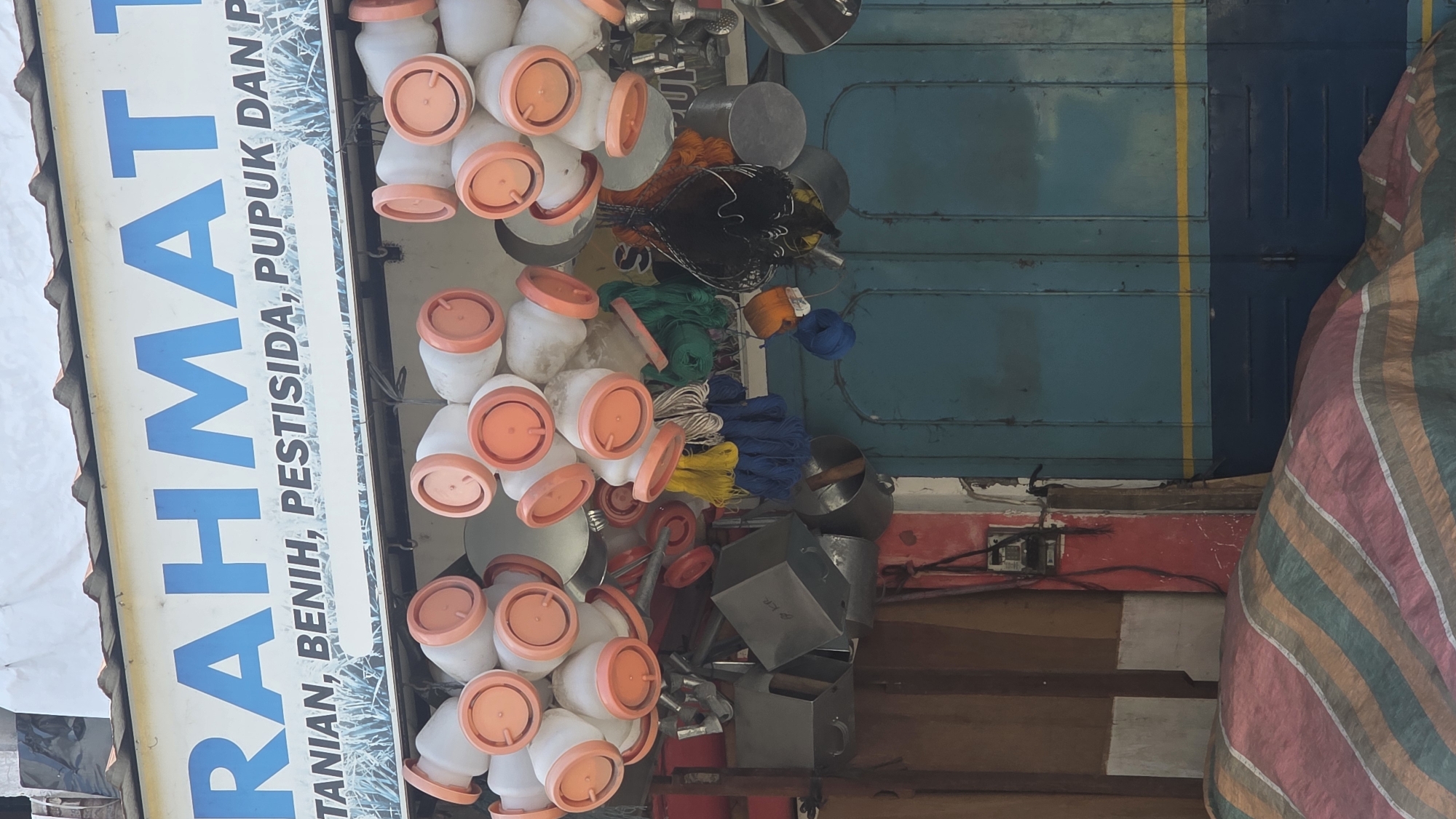 The image shows an outdoor market stall in Teungoh Baro, Indonesia. The stall displays agricultural supplies. Many small, white containers with orange lids are stacked and hung near the storefront sign, which is visible on the left edge. The sign reads "RAHMAT" in large blue letters, followed by "TANIAN, BENIH, PESTISIDA, PUPUK DAN P" in smaller blue letters. Behind the hanging containers, there's a collection of various items, including metal pots, coils of colorful rope, and other metallic equipment. The backdrop of the stall includes a teal blue door with a vertical yellow stripe and a striped tarp on the right side. Electrical wiring is visible along a red and white wall beneath the door. The stall is cluttered, but generally appears to be organized for sales. No people are visible in the image.