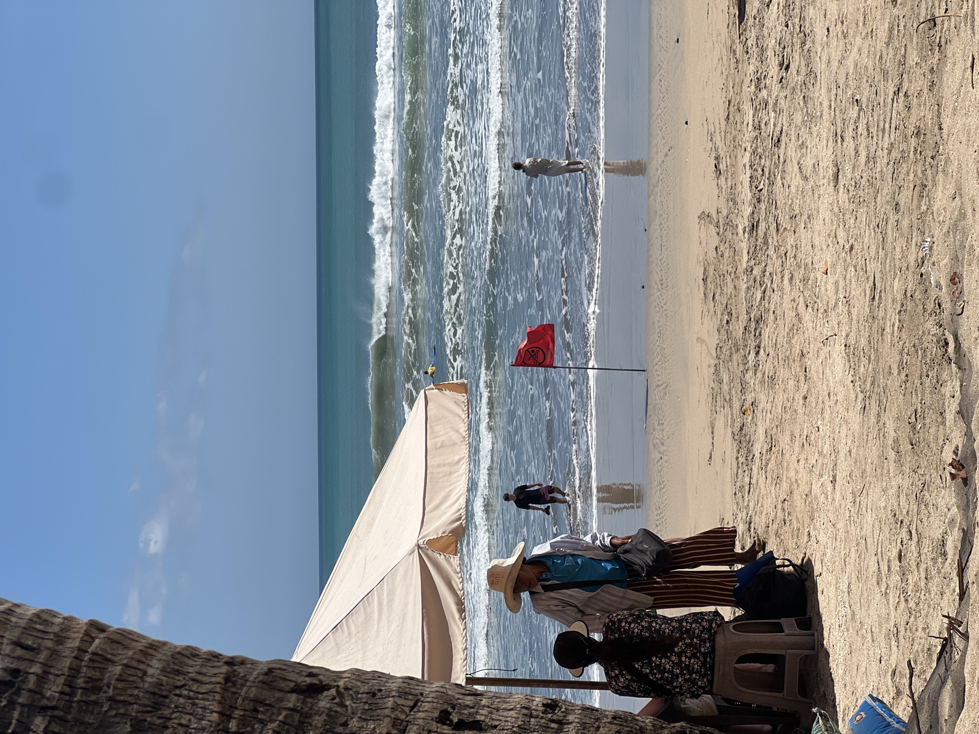 In Kuta, Indonesia, a beach scene unfolds under a clear blue sky. A large, light-colored umbrella provides shade on the sandy shore, where two individuals are seated. One person, wearing a wide-brimmed hat, a blue shirt, and striped pants, stands near the umbrella. Another person, with dark hair, sits on a small stool facing away from the camera.
The ocean, with its turquoise waters and white-capped waves, stretches to the horizon. Several surfers are visible in the distance, enjoying the surf. A red flag, likely a warning to swimmers, is planted in the shallow water. Another individual stands in the water near the red flag, facing the ocean.
The foreground of the image shows the sandy beach, with some debris and small branches scattered across it. The base of a palm tree is visible on the left side of the frame, partially obscuring the view. Bags are placed on the sand near the seated individuals. The overall atmosphere is one of a sunny day at the beach, with people enjoying the ocean and the coastal environment.