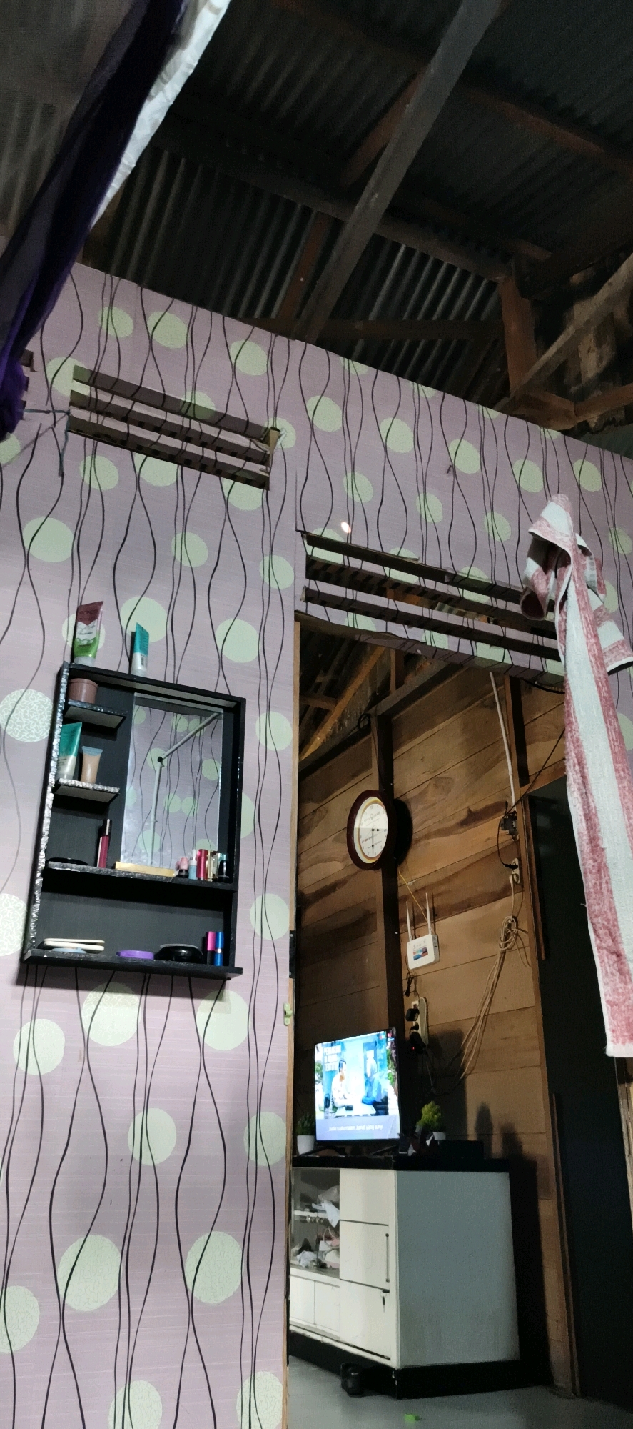 This is a vertical, eye-level, medium shot of the interior of a room in Muaro Jambi, Indonesia. The room appears to be sparsely furnished and has a rustic aesthetic. The walls are covered with a patterned wallpaper featuring light green circles and black wavy lines. In the upper portion of the image, a corrugated metal roof is visible, supported by wooden beams.
On the left side of the image, a black wall-mounted shelf unit with a mirror is attached to the wallpapered wall. The shelf unit contains several toiletries, including tubes of lotion or cream, and makeup items like lipstick and compact powder. Two horizontal vents are visible on the wall above the shelf.
To the right, the wall transitions to exposed wooden planks. A circular clock with a white face and black hands is mounted on the wooden wall. Below the clock, a white Wi-Fi router is attached to the wall with cables running down.
Further to the right, a television screen is visible, displaying what appears to be a television program. The television is placed on top of a white cabinet with black trim. Two small potted plants are positioned on either side of the television. A folded towel with red and white stripes hangs from a hook on the right side of the image, near the doorway which is not fully visible. The overall impression is of a simple living space.