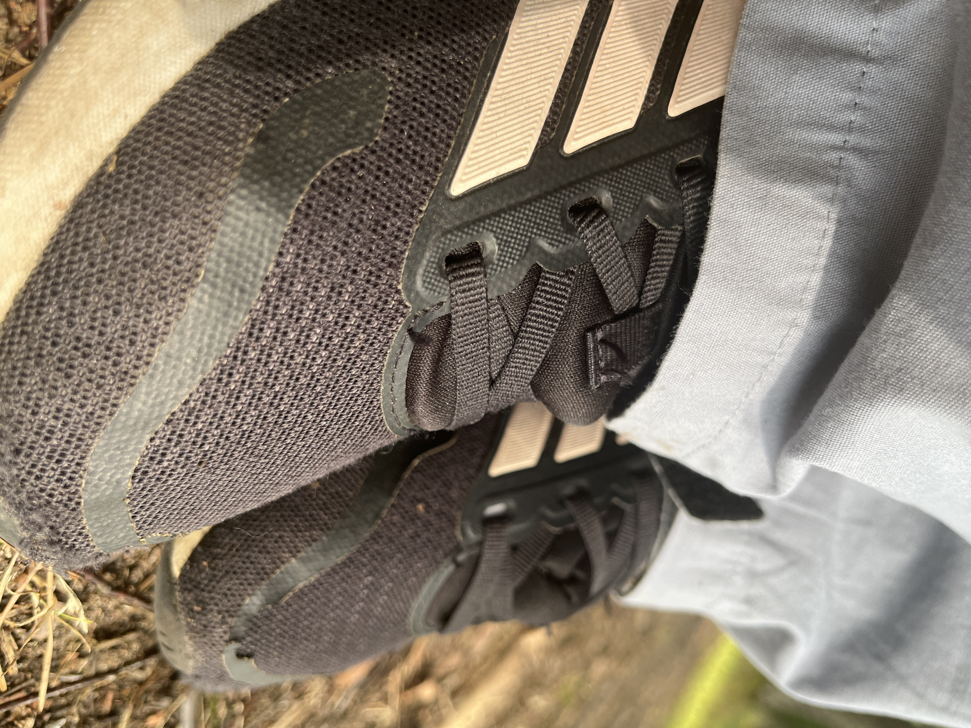 The image shows a close-up view of a person's feet wearing black Adidas sneakers with light pink stripes. The person is wearing light blue pants. The shoes are on the ground, which appears to be covered with dry grass and twigs, suggesting an outdoor environment. The lighting indicates it is daytime. The scene is devoid of any specific urban landmarks or context that would place it in Pillapalu, Estonia.