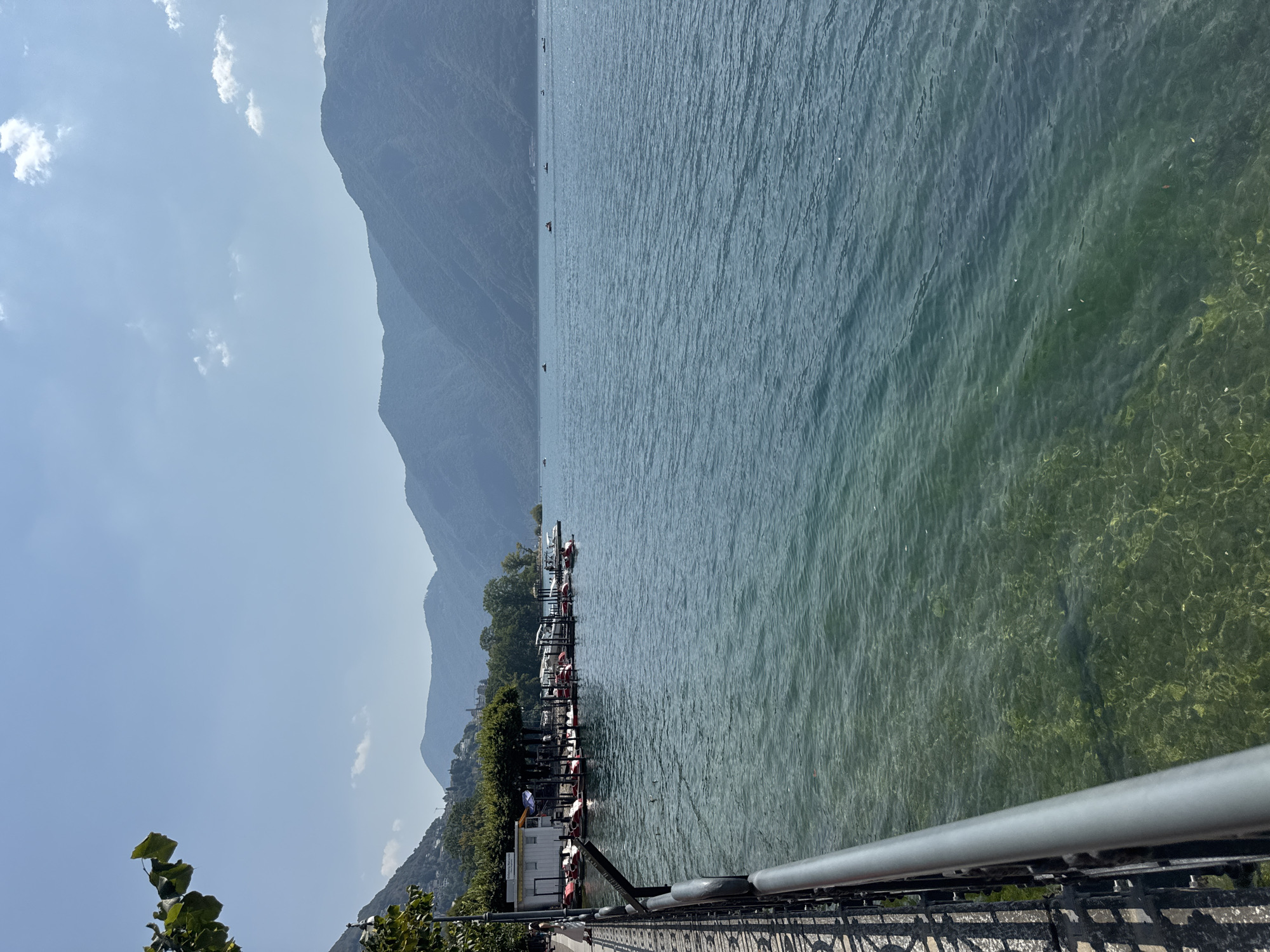 The image shows a clear, bright day at the lakefront in Lugano, Switzerland. The foreground features a paved walkway with a black metal railing. The railing's ornate design casts shadows on the walkway. To the right of the walkway is the clear, blue water of Lake Lugano. The water is shallow enough in places to see the rocky lakebed and patches of green algae. Small ripples disturb the surface of the lake.
Along the shoreline, a row of paddle boats and small recreational boats are docked. A white building with a blue awning is visible behind the boats, with several trees providing shade. In the distance, a woman in a white dress stands on the walkway. Across the lake, the hazy, tree-covered slopes of mountains rise, forming a picturesque backdrop against the bright blue sky. A few scattered white clouds drift in the sky. Several small boats are visible on the lake in the distance.