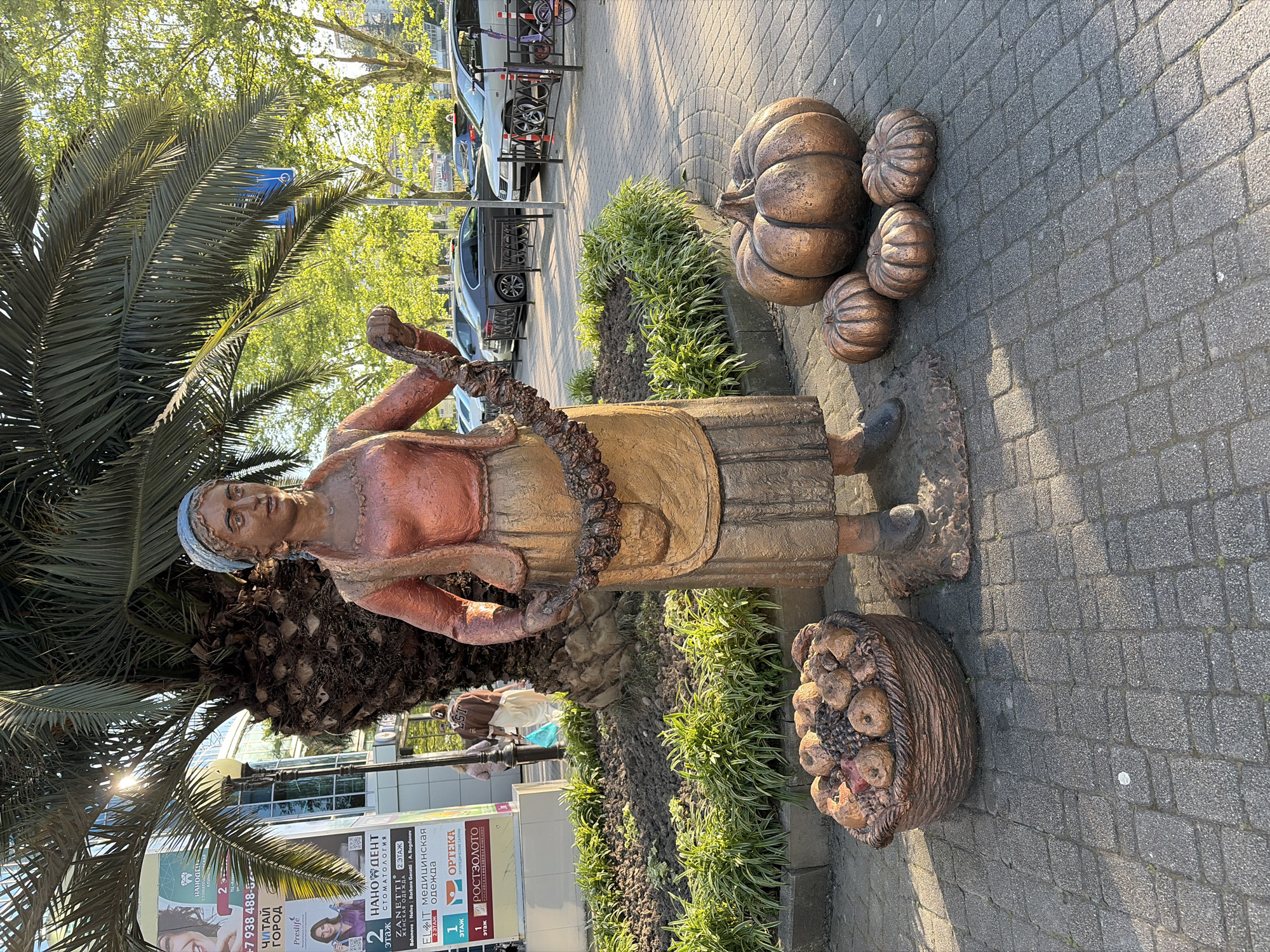 This detailed photograph captures a bronze-colored statue of a woman in traditional attire, situated on an interlocking brick pavement in a public urban setting in Sochi, Russia. The statue depicts a woman wearing a blue-grey headscarf, a reddish-orange collared shirt, a brownish-orange vest, a long brownish-gold skirt, and a lighter brownish-gold apron. She holds a garland of dark, round objects, possibly dried fruits or flowers, looped between her hands.
To the statue's left, a bronze-colored basket, sculpted to resemble woven material, is filled with various fruits, predominantly apples and grapes. To her right, a large bronze-colored pumpkin is positioned on the pavement, accompanied by three smaller bronze-colored pumpkins.
Behind the statue, a planted garden bed with green foliage and dark soil separates the paved area from a large palm tree with a thick, textured trunk and extensive fronds. Sunlight streaks through the upper left section of the palm fronds. In the mid-ground, a modern multi-story building with glass windows is visible, adorned with large advertisements in Russian. Identifiable text includes "НАНОДЕНТ СТОМАТОЛОГИЯ" (NANODENT STOMATOLOGY), "ZANETTI ЖЕНСКАЯ ОДЕЖДА" (ZANETTI WOMEN'S CLOTHING), "ELIT МЕДИЦИНСКАЯ ОДЕЖДА" (ELIT MEDICAL CLOTHING), and "РОСТЗОЛОТО" (ROSTZOLOTO), along with floor indicators like "2 ЭТАЖ" (2ND FLOOR) and "1 ЭТАЖ" (1ST FLOOR).
Several blurry individuals are visible walking in the background near the building. Further back, a street or parking area contains multiple parked cars, including sedans and SUVs, separated by a black metal fence. A child's scooter is partially visible next to the fence on the right. Lush green deciduous trees line the distant background. The scene is illuminated by natural sunlight, casting shadows to the right, suggesting either morning or late afternoon.