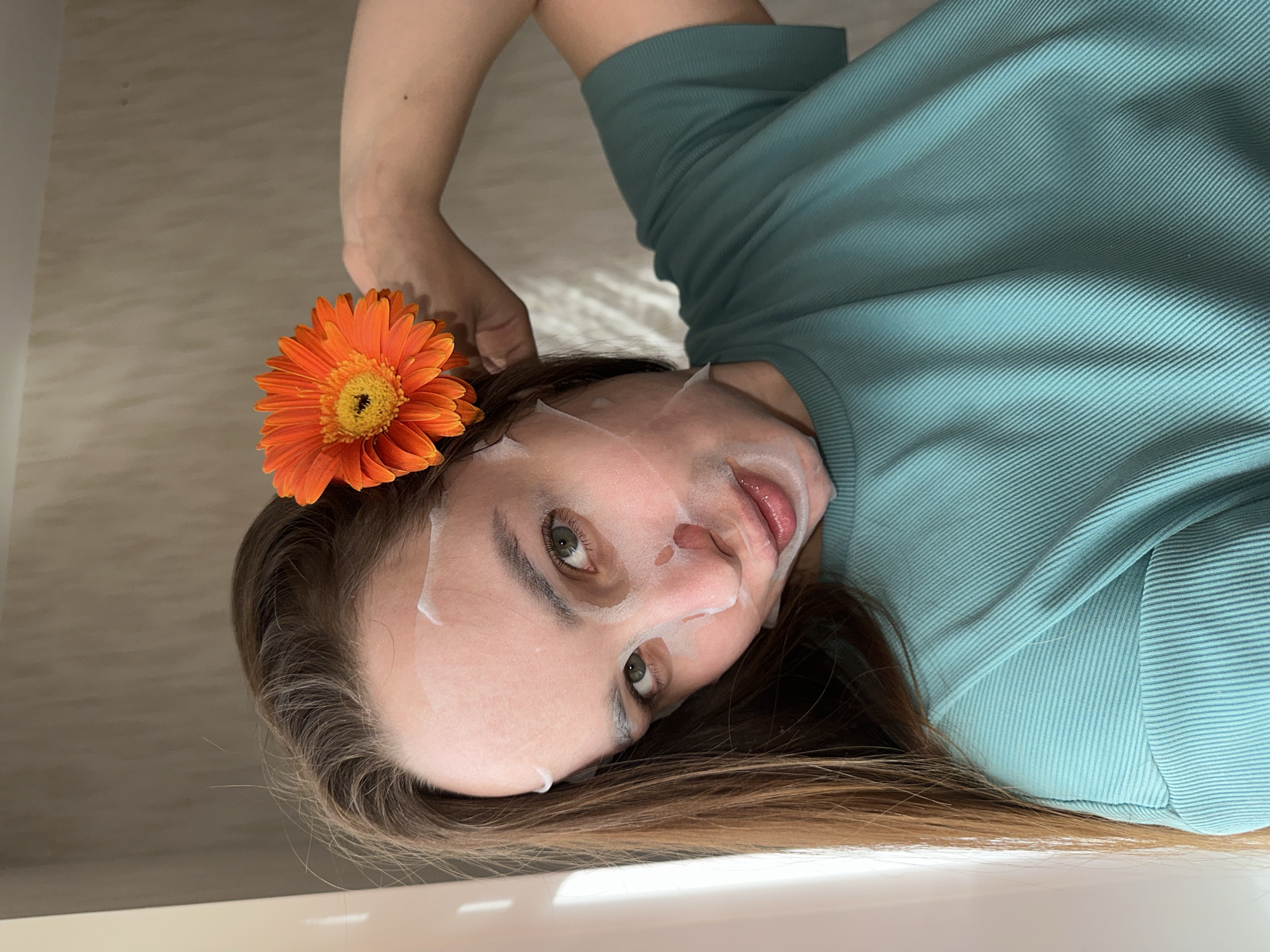 An image, captured in Chaikovsky, Russia, depicts a person lying down indoors, engaged in a self-care routine. The subject, likely female, has light brown hair fanned out around their head and is wearing a transparent facial sheet mask covering most of their face, with cutouts allowing their eyes, nose, and mouth to be visible. Their eyes are open and gaze directly at the viewer. A vibrant orange gerbera daisy flower is placed near their temple, resting on their hair.
The person is wearing a long-sleeved top in a teal or mint green color, characterized by a distinct ribbed or striped texture in the fabric. They appear to be resting on a smooth, light-colored surface in the foreground, possibly the edge of a bathtub or a bed frame. The background is a plain, light-toned wall or surface. The lighting is soft and diffused, creating a serene and relaxed atmosphere. No visible text is present in the image. The overall impression is one of relaxation and personal indulgence.