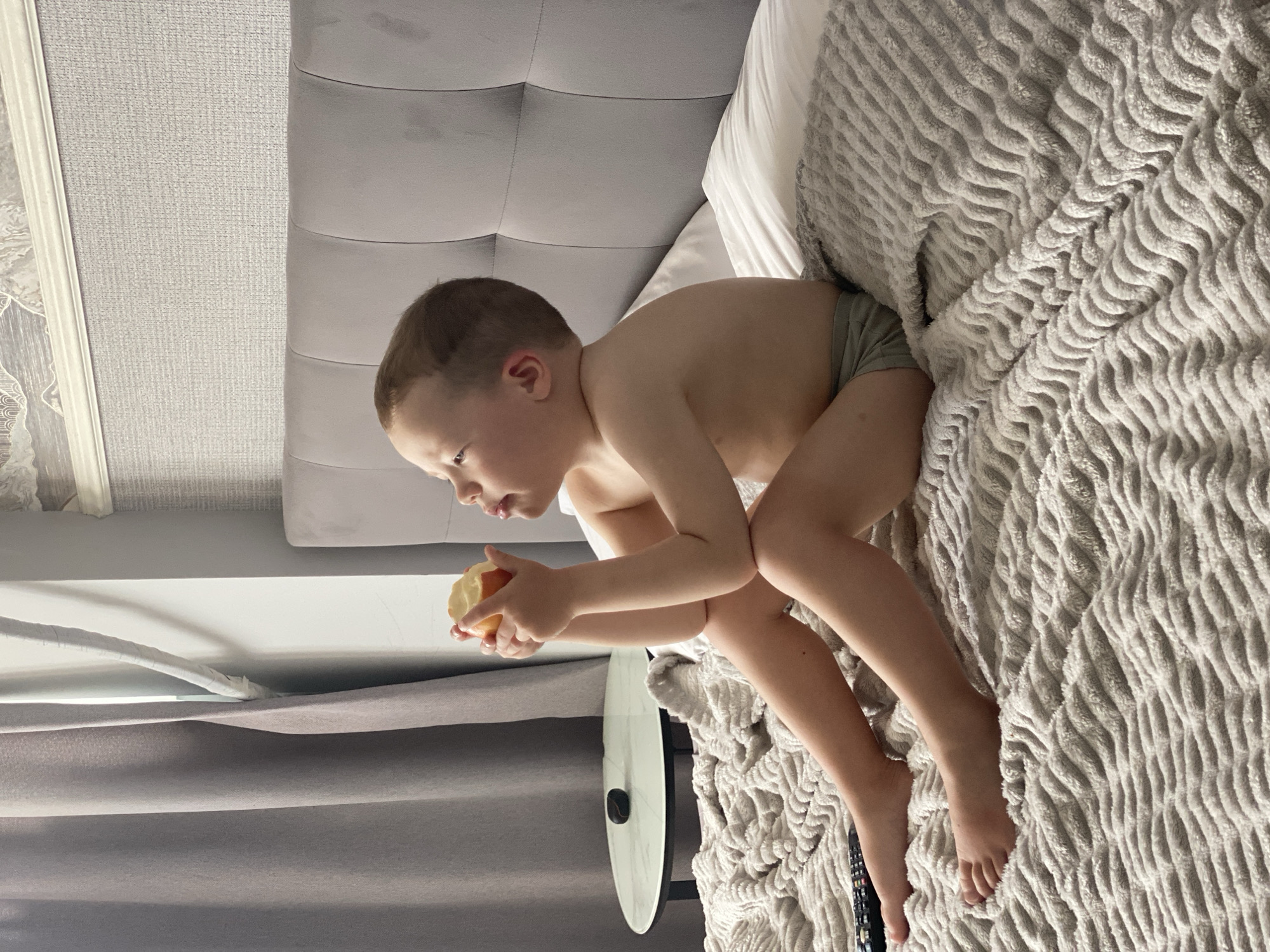 A young boy, likely between 3-5 years old, sits on a bed and holds an apple. He is wearing grey underwear. The bed has a grey tufted headboard and is covered with a textured grey blanket. A white pillow is visible behind the boy. The room appears to be a bedroom, and the lighting suggests it could be daytime. The boy is looking down at the apple he is holding, and his mouth is slightly open, as if he is about to take a bite. The overall impression is one of a candid moment of a child enjoying a snack. The background includes a grey wall with a textured wallpaper and a white trim. There is also a glimpse of a window or curtain on the left side.