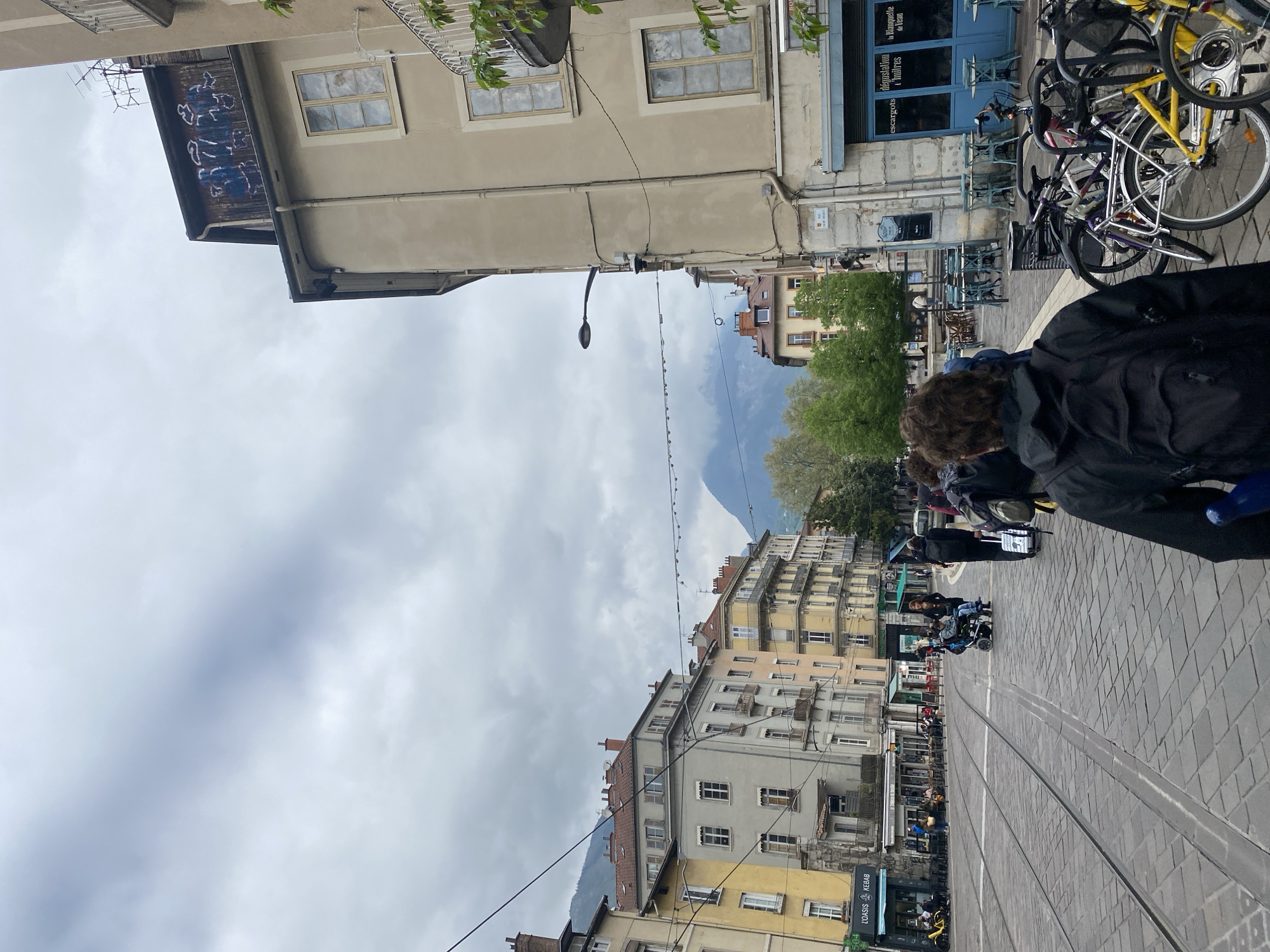 The image depicts a street scene in Grenoble, France, characterized by tall buildings, tram tracks, and a mountainous backdrop. The architecture features classic European facades with multiple windows and varying shades of yellow and gray. Tram tracks run through the paved street, indicating a central transportation route. Pedestrians are visible, including a person in a black jacket and a possible wheelchair user. Bicycles are parked, with one prominently yellow bike among others. Overhead cables and a street lamp cross the sky. Storefronts with signs, possibly for food establishments like "LOASIS KEBAB", add to the urban setting. The sky is overcast, suggesting a cloudy day. The mountains in the background provide a natural contrast to the urban environment.