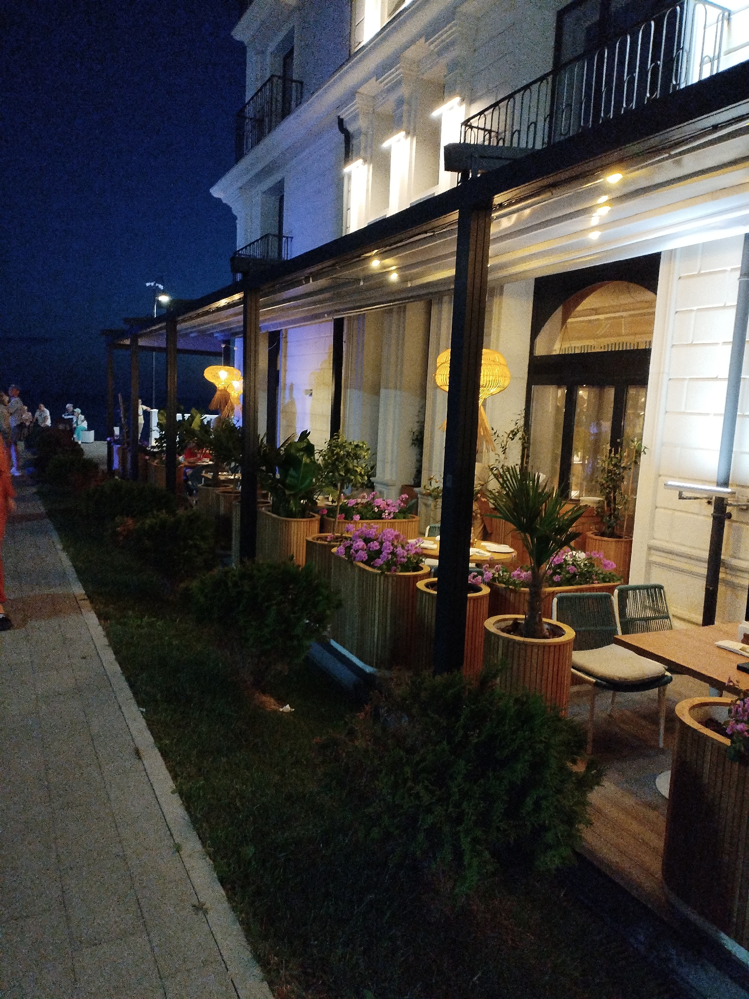The image depicts a nighttime view of an outdoor seating area of a restaurant or cafe, likely located in Vinogradny, Russia. The scene is illuminated by warm, artificial lighting from string lights and pendant lamps. The architecture of the building features white walls with ornate detailing and balconies. In the foreground, a paved walkway runs alongside a strip of dark green grass and several large, wooden planter boxes filled with lush green plants and vibrant purple flowers. Beyond the planters, the seating area is visible, with wooden tables and chairs, some of which are occupied by patrons. In the background, more people can be seen standing and socializing on a walkway or promenade, with a dimly lit body of water, possibly the Black Sea, visible in the far distance under a dark blue sky.