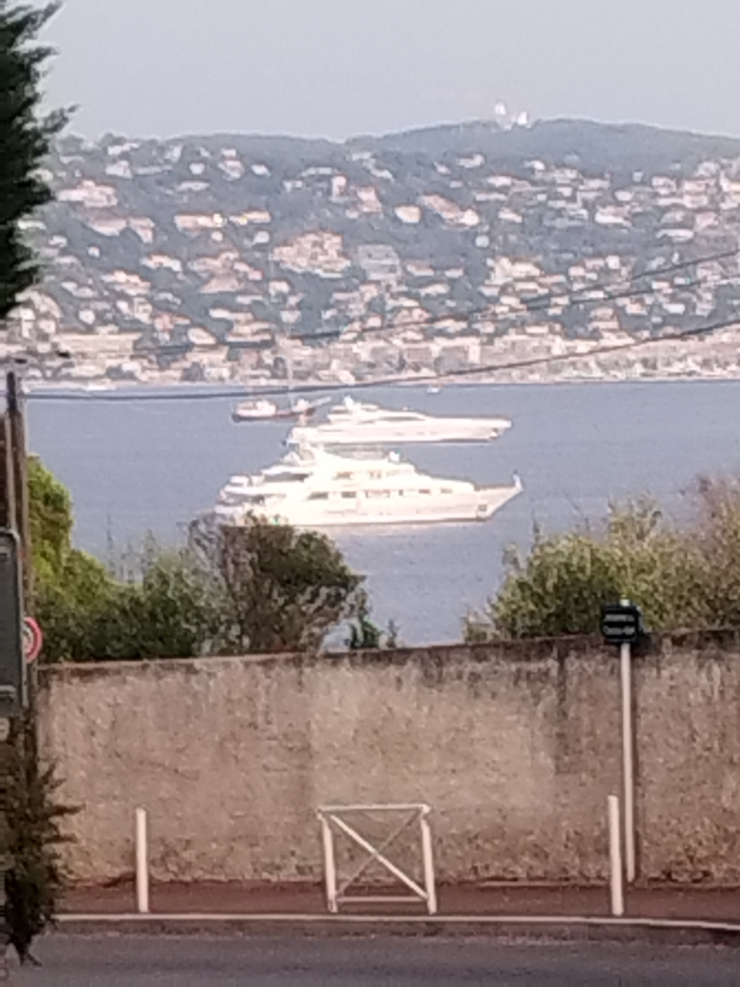 The image shows a waterfront scene with a city built on a hillside in the background. In the foreground, there is a low, textured concrete wall. Behind the wall, there is a body of water where two large white yachts are docked. One yacht is larger than the other and is positioned closer to the shore. The city in the background is densely populated with many buildings visible on the slope of the hill. The sky is overcast, and the lighting suggests it might be late afternoon or early evening. The scene appears to be a view from a slightly elevated position looking down towards the water and the city.