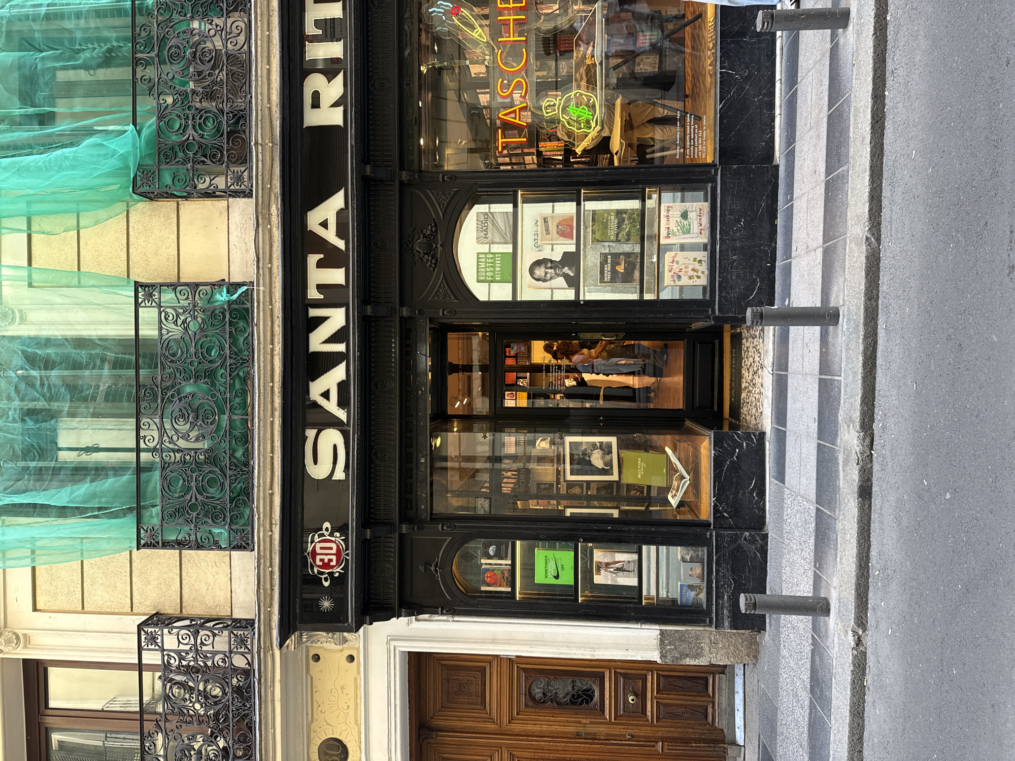 The image shows the facade of the Taschen store on Santa Rita Street in Madrid, Spain. The store is a small, elegant building with a black and marble front. The name "SANTA RITA" is prominently displayed in white letters above the entrance, and the Taschen logo, including a neon sign, is visible in the window. A window displays books and a sign. The door is made of wood and glass. A green construction tarp covers windows above. People are seen inside the store. The street is paved and has bollards along the edge.
