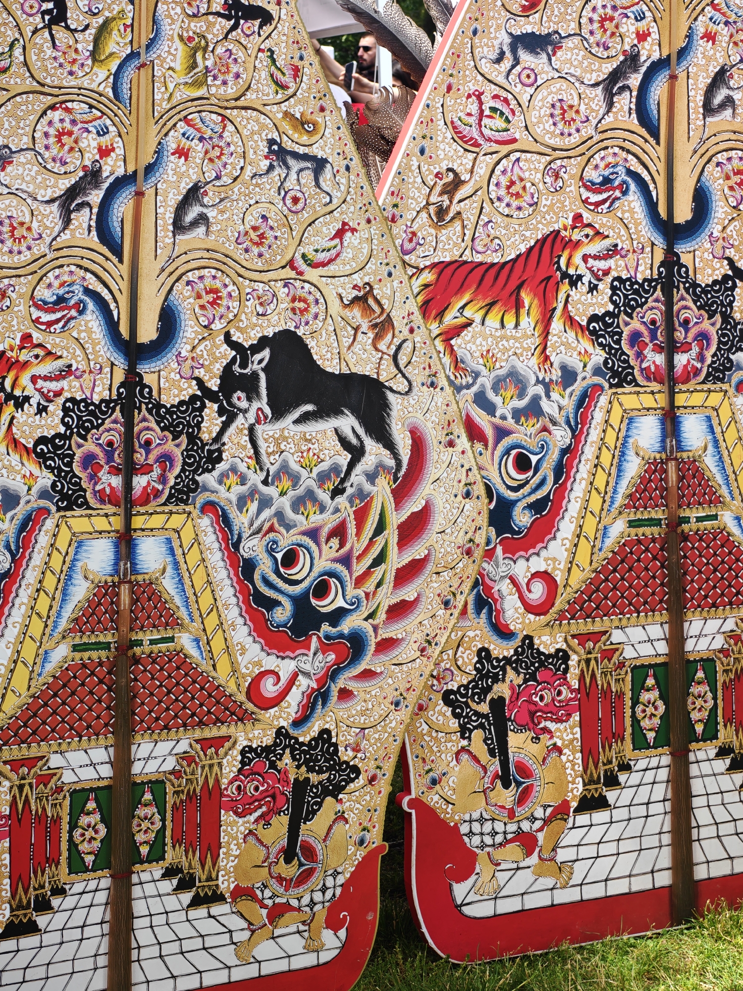 The image displays two ornate, traditionally crafted puppets, possibly from a Southeast Asian cultural tradition, positioned outdoors on green grass. The puppets are intricately painted with gold, red, blue, and black details, featuring mythical creatures like dragons and tigers, as well as human-like figures and architectural elements resembling temples. The puppets are made of wood or similar material and have wooden poles running vertically through them, suggesting they are designed to be manipulated. In the background, out of focus, are parts of what appear to be other decorative elements, possibly more puppets or costumes, and a person's head and shoulders, seen from the side, wearing sunglasses. The setting appears to be a cultural event or festival. This scene was captured in Sofia, Bulgaria, as part of an exhibition.