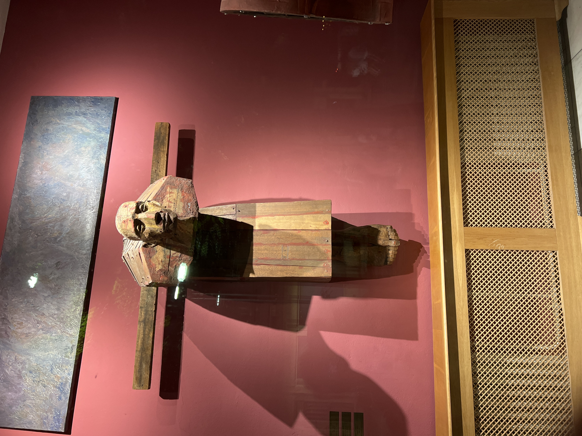 The image displays a wooden sculpture of a man in stocks, mounted on a deep red wall. The sculpture depicts a man's head and upper torso, with his face carved in a pained expression, set within a wooden collar. The body is a wooden post with foot shackles at the bottom. A large, abstract painting hangs above the sculpture. In the foreground, a wooden radiator cover with a lattice pattern is visible. The sculpture and artwork appear to be displayed in a museum or gallery setting. The image was taken in the Constanța Museum of National History and Archaeology in Constanța, Romania.