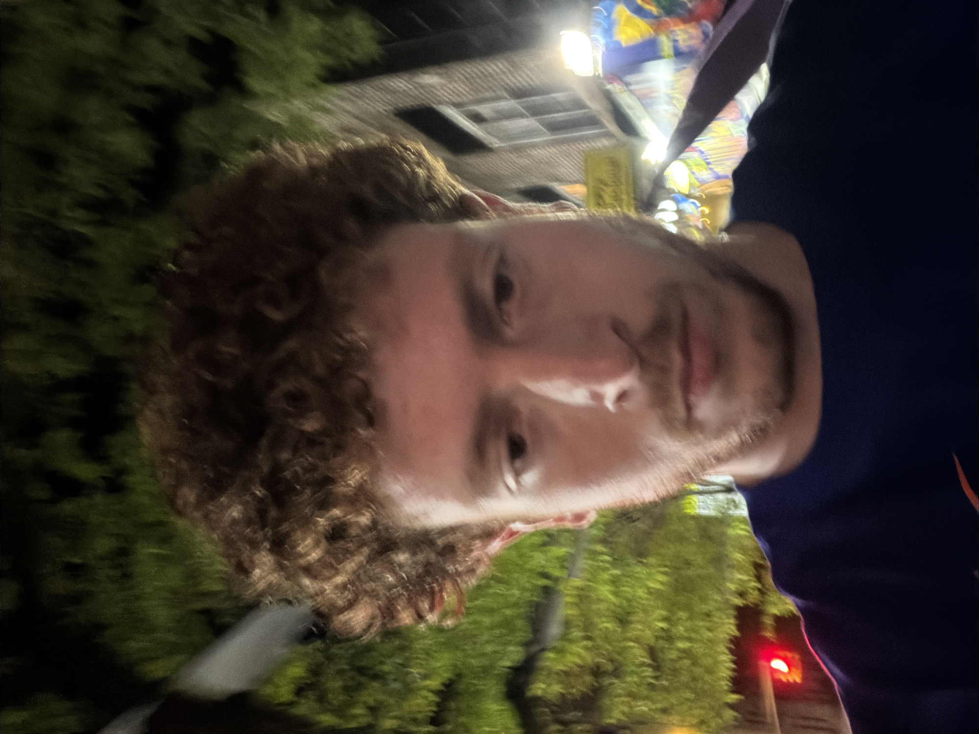 A selfie of a white male with curly brown hair and a beard. He is wearing a dark blue shirt. The background appears to be a street scene at night, with trees, streetlights, and a building visible. The location is likely in the city of New York, United States of America, based on the general appearance of the urban environment.