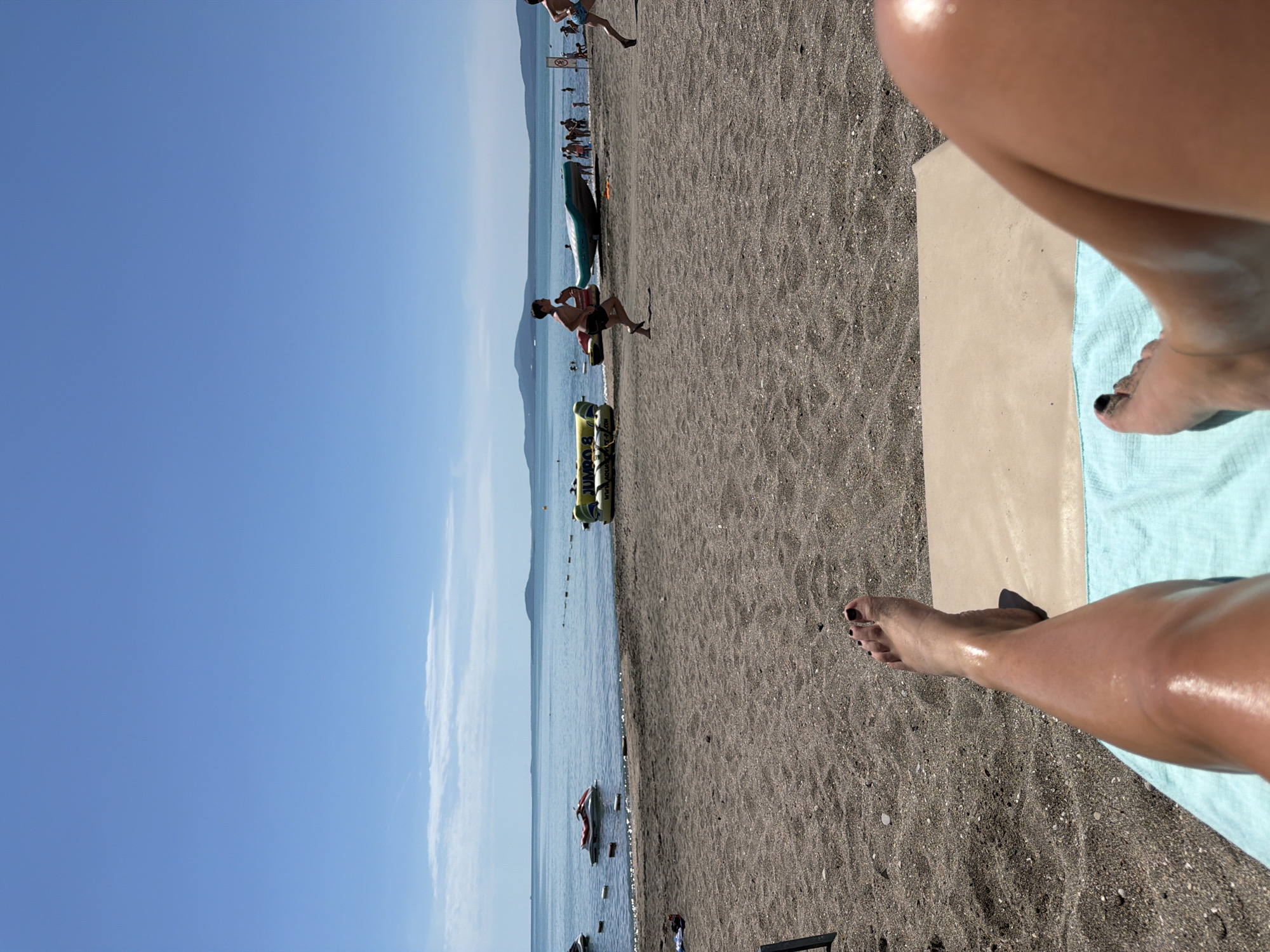 The image depicts a beach scene on a sunny day, likely in Burgas, Bulgaria. In the foreground, a person's legs and feet are visible, resting on a light-colored mat and a blue towel. The person's legs are tanned and appear to have oil or lotion on them, reflecting the sunlight. The beach itself is sandy with dark pebbles.
In the middle ground, the sea is calm and blue, with several objects floating on its surface. To the left, two jet skis are partially visible, one red and white, and another dark. Further out, a yellow inflatable water toy, labeled "JUMBO 8", is moored. Several people are scattered along the shoreline, with some wading in the water and others walking on the sand. Two young boys are running on the beach, one in the mid-ground and another further to the right.
In the background, a clear blue sky stretches across the horizon, with a few wispy white clouds. Distant landmasses, appearing as hills or mountains, are visible across the sea. The overall impression is of a relaxing and active day at the beach.