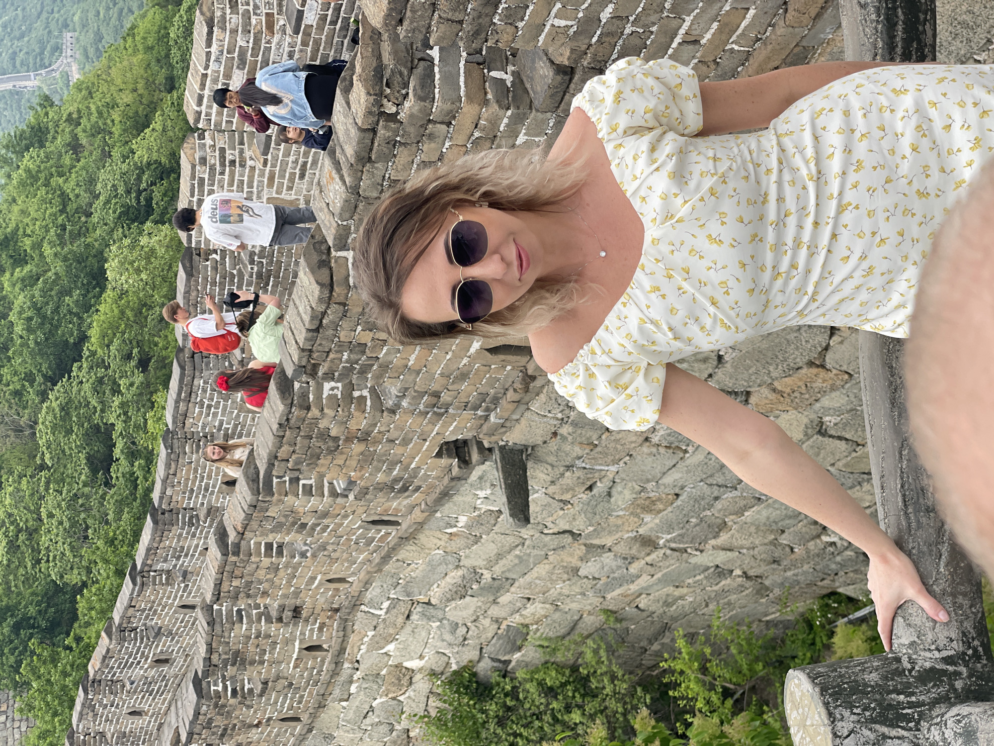 The image shows a woman with sunglasses and a floral dress posing on the Great Wall of China in the Huairou District. Several other tourists are walking in the background along the wall. Lush green trees can be seen in the valley below the wall. The Great Wall itself is constructed of grey bricks.