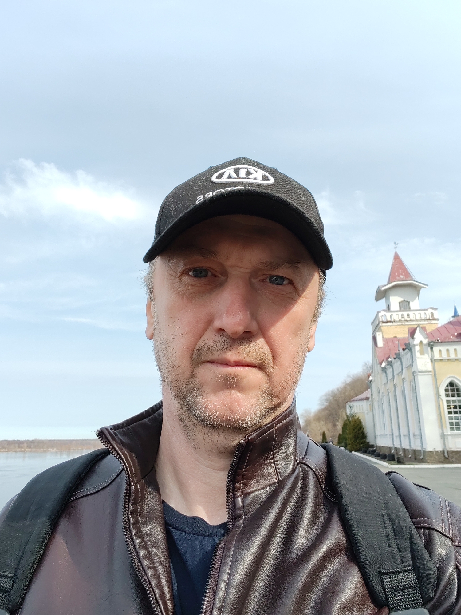 A medium-shot selfie shows a man wearing a black KIA baseball cap and a brown leather jacket over a blue t-shirt, with a black backpack strap visible over his left shoulder. He has short, fair hair, blue eyes, and a light beard. The background reveals a bright, cloudy sky and a building in the city of Sarapul, Russia. The architectural style of the building features white walls, rows of columns, red roofing, and a tower-like structure. The man is positioned on a paved surface that suggests a street or walkway near a body of water with trees lining the opposite shore.