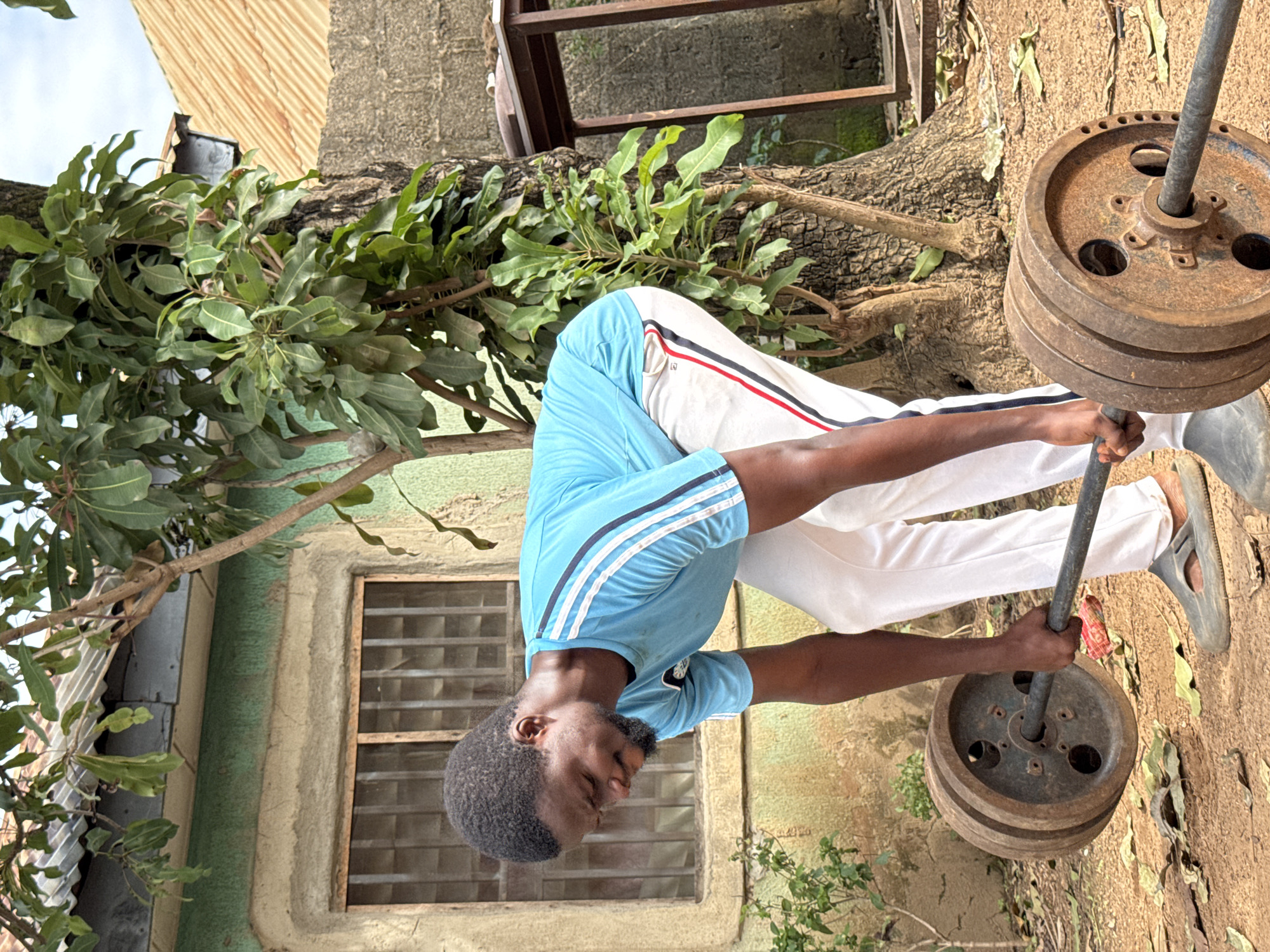 An African male is shown performing a deadlift outdoors in Akwanga, Nigeria. He is wearing a blue t-shirt with white stripes and white pants with red and blue stripes. The man is in the process of lifting a barbell loaded with weights. The environment appears to be a residential yard with a rustic building in the background, a tree, and dry ground covered with leaves. The building has a green painted wall and a window with bars. A corrugated metal roof is visible on another structure. The overall setting is informal and appears to be a home gym setup.