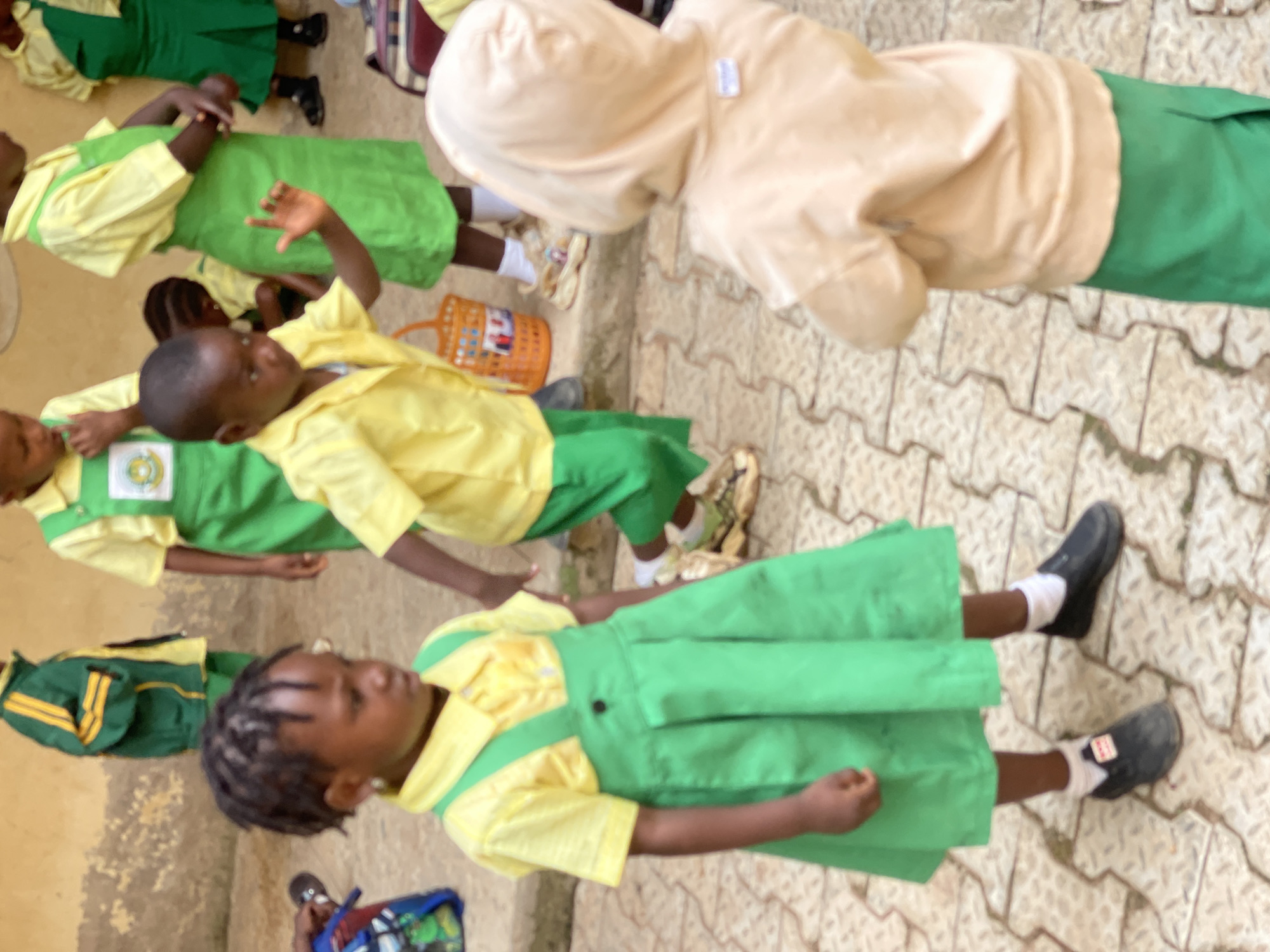The image captures a group of children in school uniforms outside what appears to be a school building in Akwanga, Nigeria. The foreground features two young girls and a boy, all wearing yellow short-sleeved collared shirts and green pleated skirts or jumpers over their shirts. The girl on the left has her hair in braids and wears small hoop earrings. She is looking down and to the left, with her right hand slightly raised. The boy in the center is facing the viewer and has his right hand raised as if to greet or signal. He is wearing a yellow shirt and green jumper. Behind him, another child in a green uniform is visible. To the right, a child wearing a light beige hooded sweatshirt and a green skirt is seen from behind, facing away from the viewer. In the background, another child in a green and yellow jacket and green pants is partially visible. The ground is paved with textured interlocking blocks. There are backpacks and a basket-like object visible on the ground, suggesting a school setting or an outdoor play area. The lighting indicates it is daytime.