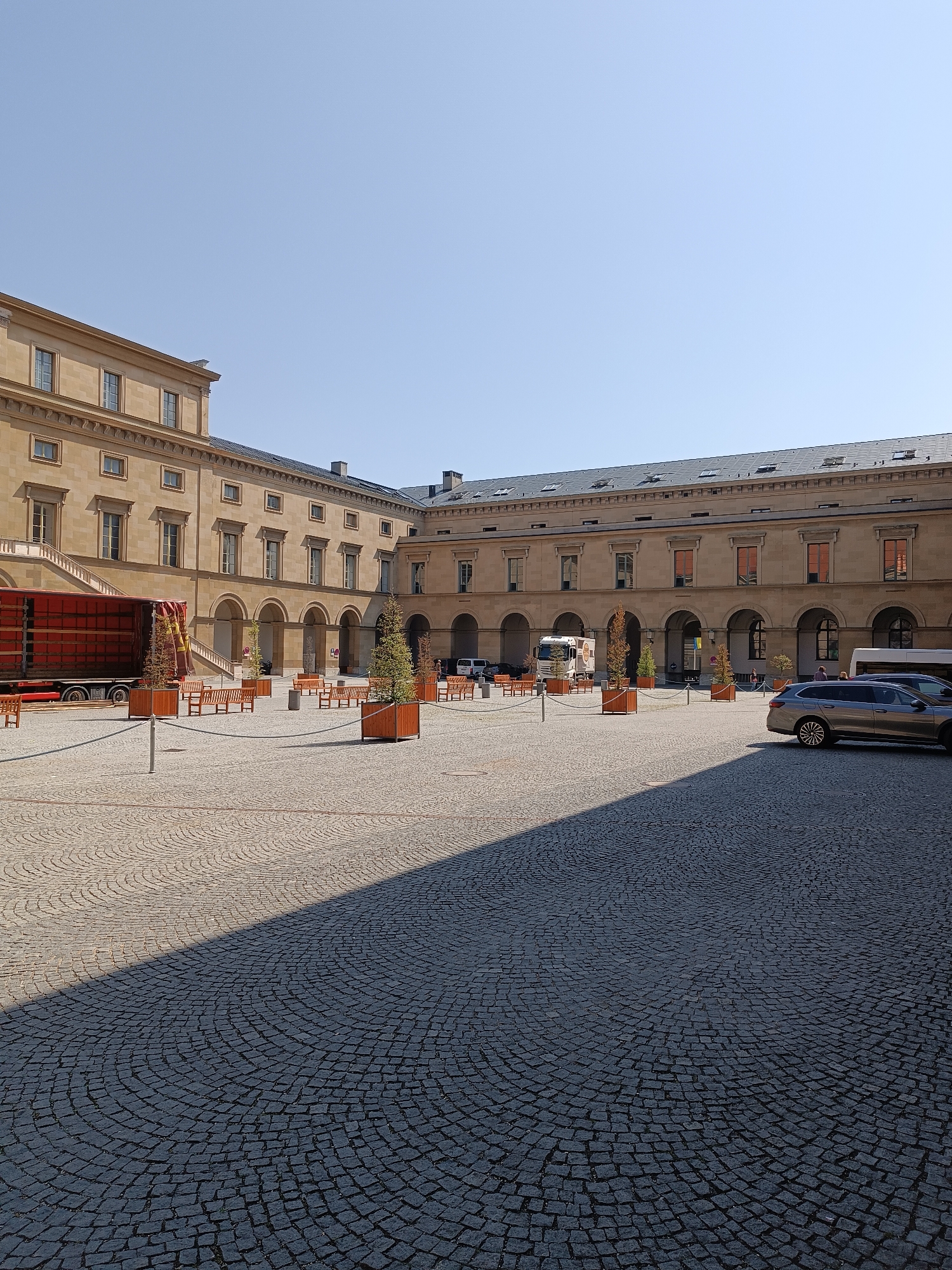 The image shows a wide, paved courtyard in Munich, Germany, likely within a historical or institutional complex given the architecture of the surrounding buildings. The buildings are of light-colored stone with classical architectural elements, featuring multiple stories, arched walkways, and numerous windows. The courtyard itself is paved with cobblestones arranged in a circular pattern.
In the foreground, the cobblestone pavement is divided by a distinct shadow, indicating a sunny day. The shadow falls across the right side of the image, creating a contrast between the sunlit and shaded areas.
Several elements are present in the courtyard. On the left side, a large red truck with a trailer is parked, partially obscured by a tree in a planter. In the center of the courtyard, several wooden benches are arranged, some in clusters and others individually. There are also several tall, narrow planters with small trees, contributing to the landscaping of the space.
In the background, near the arched walkways, a white truck is visible, and further to the right, a white bus or van is parked. A grey car is parked in the right foreground, facing away from the camera. A few people are visible in the background, walking near the arched walkways and near the parked vehicles. The sky is clear and blue, suggesting good weather conditions. The overall impression is one of a calm, public space, possibly a plaza or an inner courtyard of a museum or governmental building.