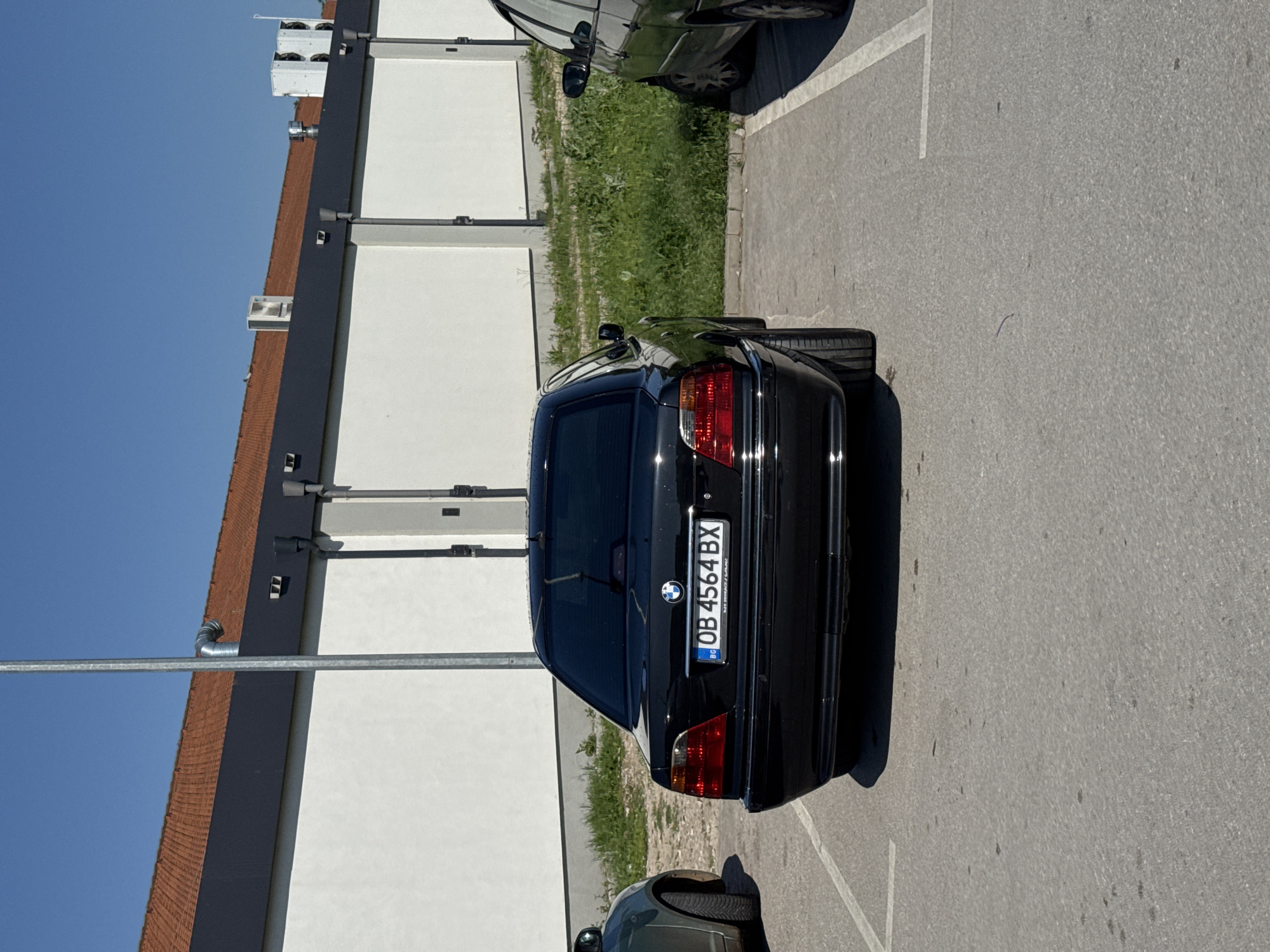 The image depicts the rear of a black BMW car parked on asphalt within an urban parking area. The license plate reads "OB 4564 BX", indicating registration in Bulgaria. Adjacent to the BMW is another car, partially visible, suggesting a full parking lot. A large building with white walls and a reddish-brown roof is in the background. Vegetation is growing alongside the building. The sky is clear and blue. The location is Lovech, Bulgaria based on the license plate code.