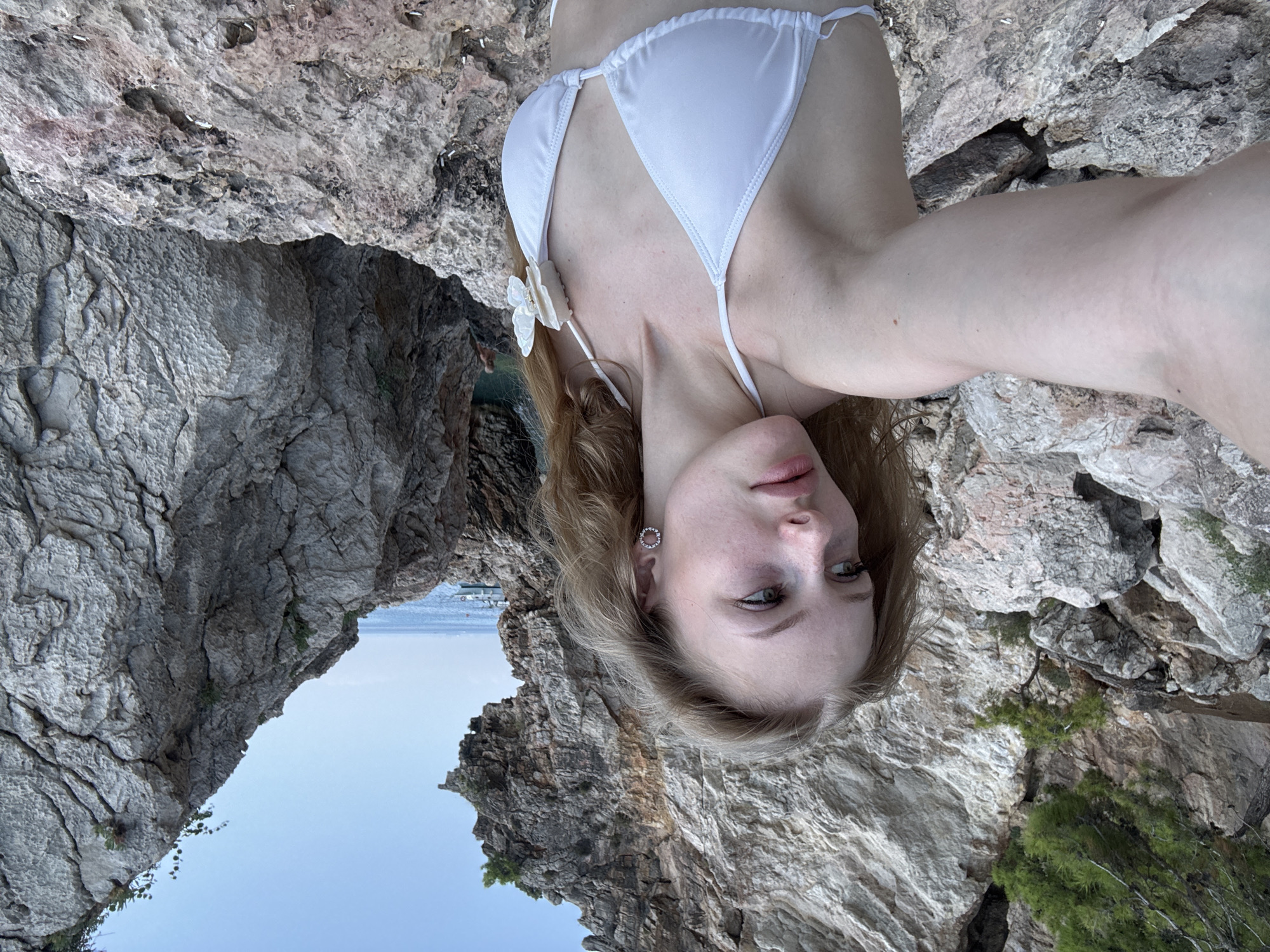 A young woman with long blonde hair, wearing a white bikini with a butterfly embellishment on the strap, stands on a rocky shore. She is taking a selfie, looking off to her left. The background features rugged, grey rock formations and the blue sea with a few distant boats. The scene appears to be a natural, coastal environment.