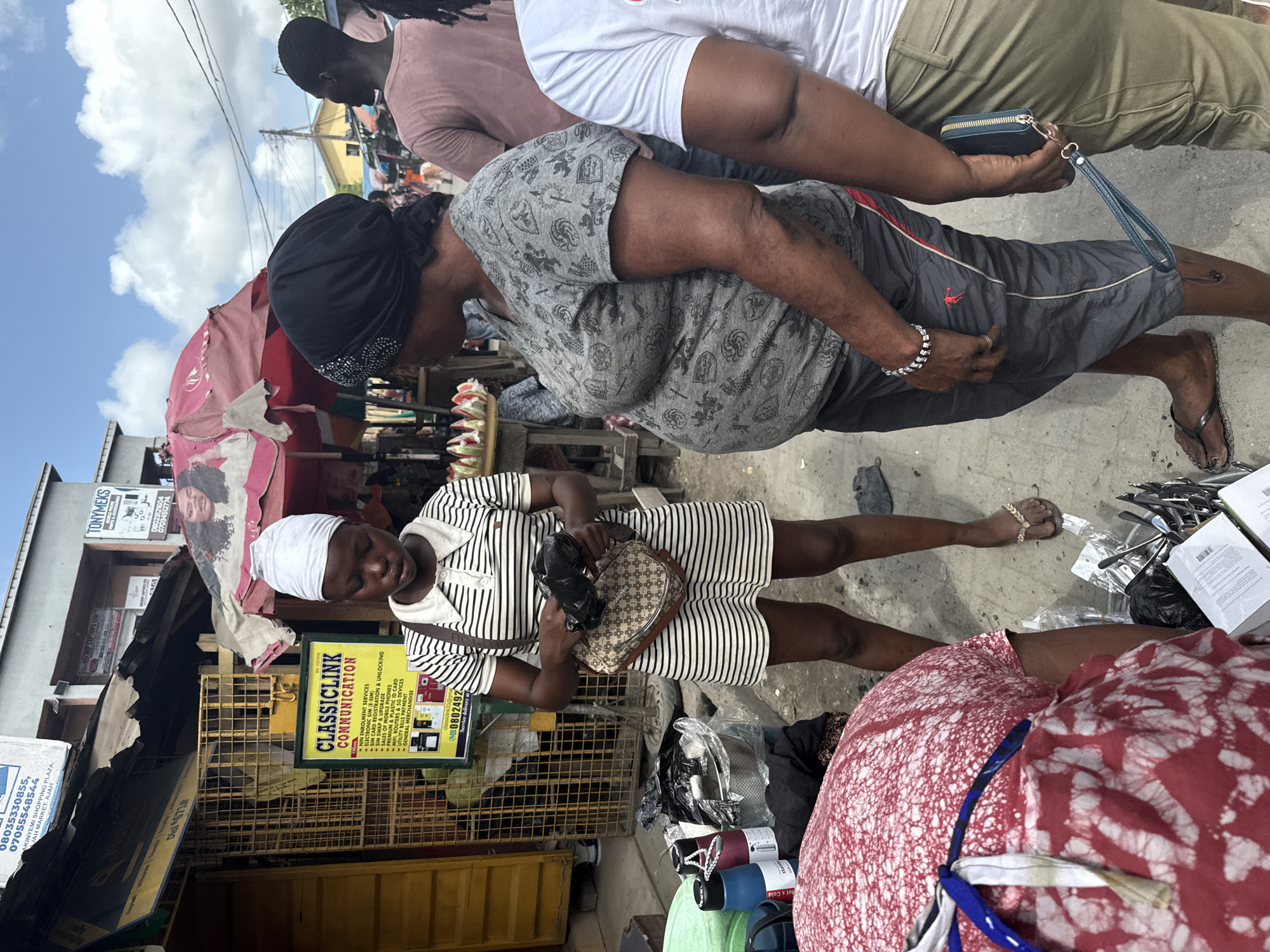 The image depicts a street scene in Lagos, Nigeria. In the foreground, a woman wearing a striped dress and a white headscarf holds a handbag, standing amidst various goods likely for sale. Behind her, a sign advertises "Classiclink Communication," providing services such as SIM card registration and phone sales. Other signs and advertisements are also visible on nearby buildings. Several individuals are walking by, including a woman in a grey patterned top. The setting is a busy marketplace, evident from the diverse assortment of goods and the presence of multiple vendors.