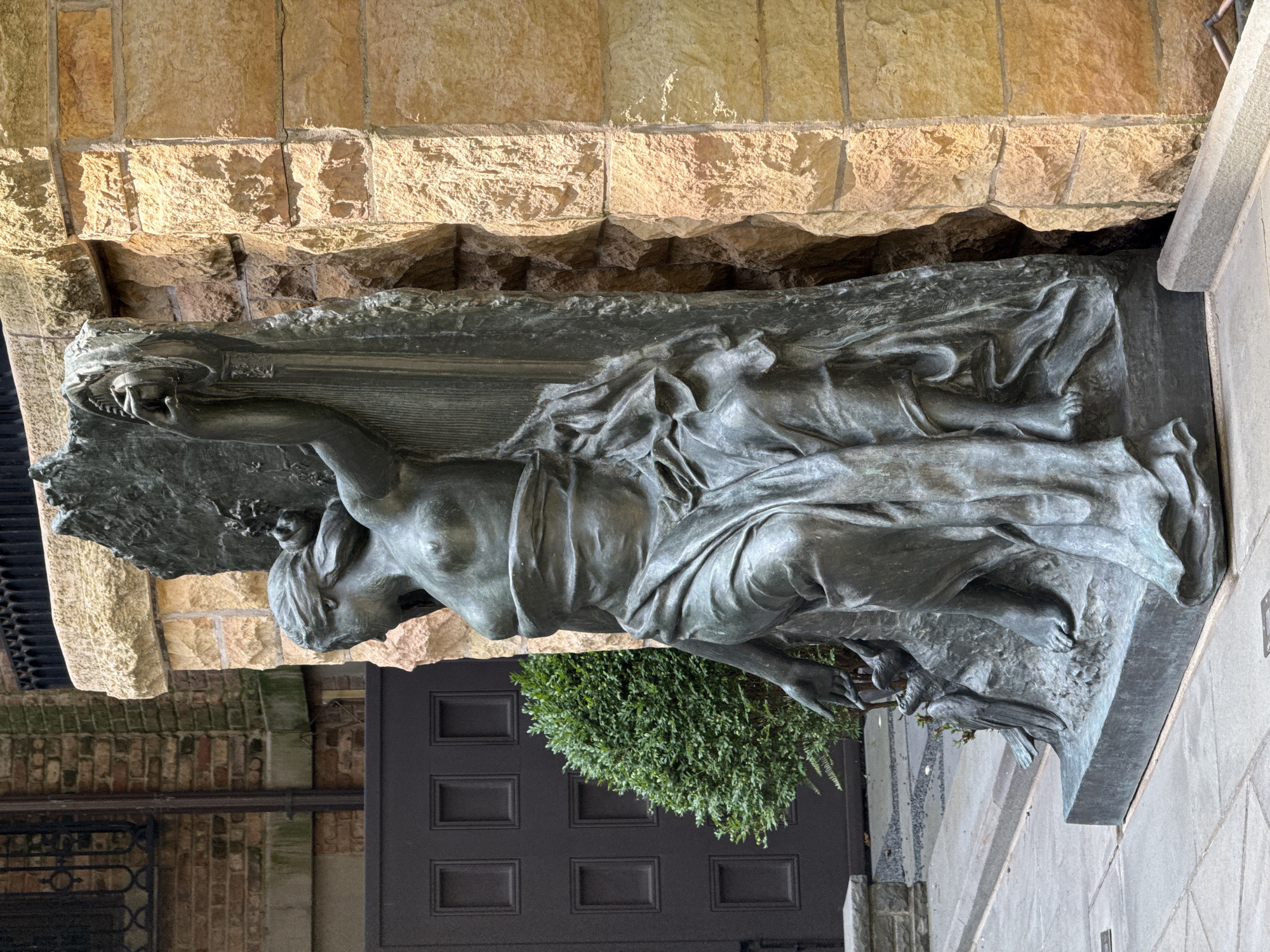 A bronze sculpture of a partially clothed woman is positioned outdoors next to a rough-hewn stone wall. The sculpture depicts the woman sitting with her left leg bent and her right leg extended. She wears a draped cloth that covers her lower body and shoulders. Her left arm is extended downward, with her hand near two birds. Her right arm is raised, holding a circular object. The sculpture is mounted on a rectangular base. Behind the sculpture, a dark brown double door with square panels is visible, as is a neatly trimmed green shrub. The background includes a brick building with a black metal railing and a wall constructed of large, irregular light brown stones. The scene is likely in a public area, possibly a garden or courtyard, in Chicago, Illinois, United States of America.