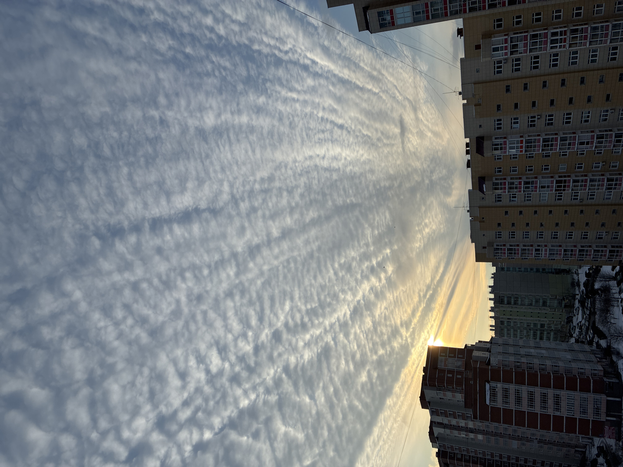 The image captures a view between buildings in an urban setting. Apartment buildings with multiple windows are visible on either side of the frame. The sky above is filled with a layer of striated clouds. Sunlight appears to be filtering through the clouds near the horizon, creating a bright glow. The environment is quiet. No people are visible.