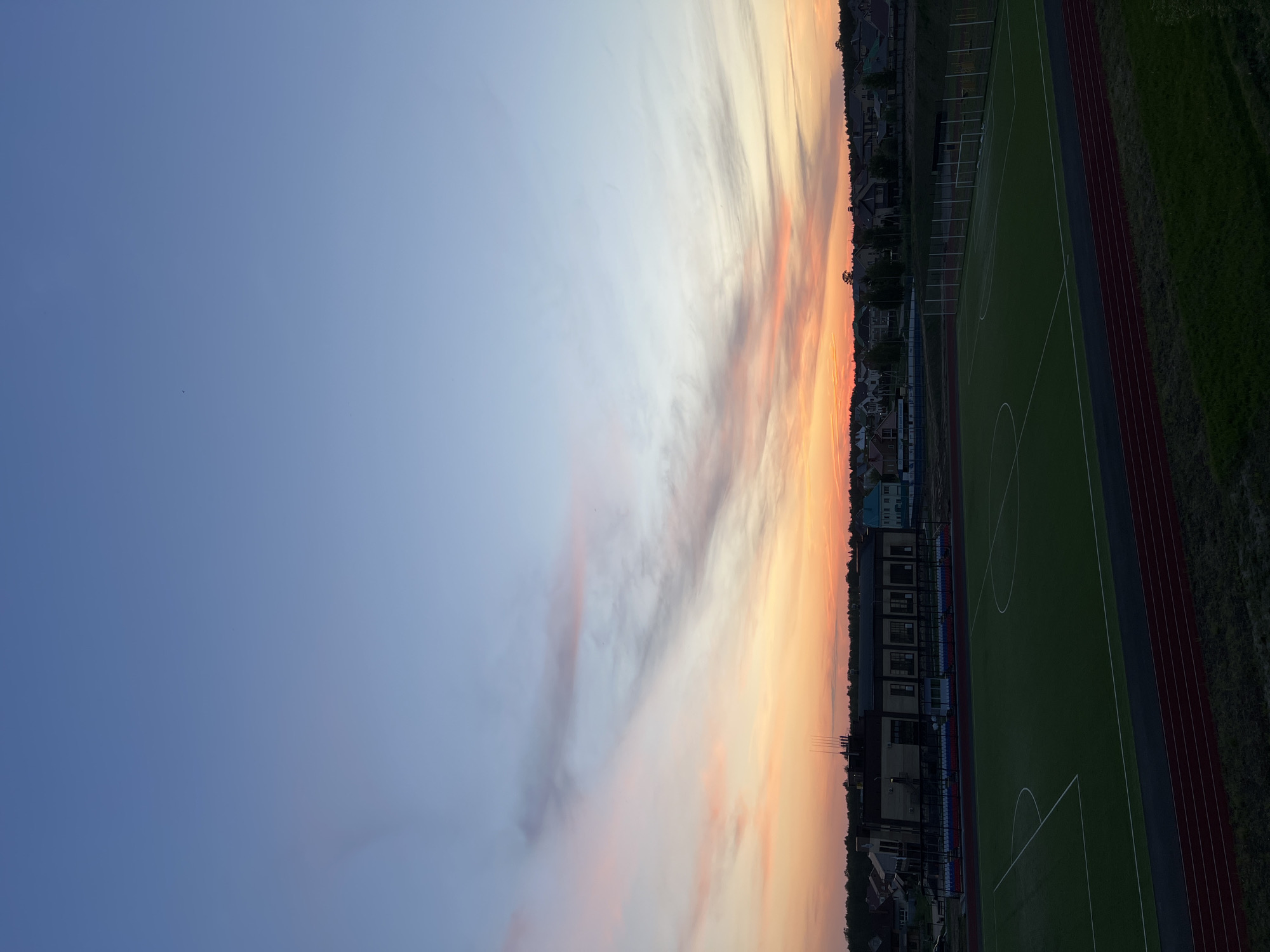 The image shows a view of a soccer field in Noviy, Russia at sunset. The sky is filled with orange and pink clouds fading into a blue gradient. Adjacent to the soccer field, there is a running track. Buildings are visible in the background, located near the horizon.