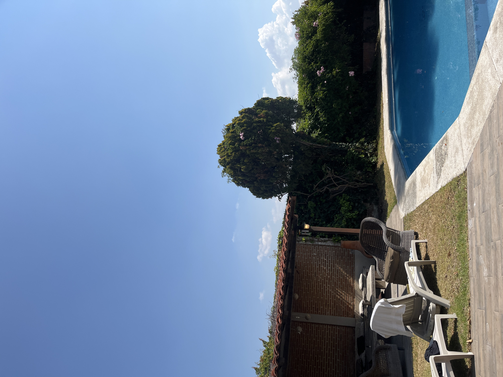 The image depicts a tranquil scene in El Oasis Valsequillo, Mexico, featuring a clear blue sky with scattered clouds above a residential property. The property includes a swimming pool with bright blue water, bordered by a concrete deck and adjacent green foliage. The scene incorporates outdoor furniture, including wicker and plastic chairs, partially situated on a grassy lawn that transitions into a tiled patio. A brick structure with a red tile roof sits to the left of the chairs. Lush greenery, including a large tree with pink blossoms, is present along the property line, adding a sense of privacy and seclusion to the space. No people are visible in the scene.