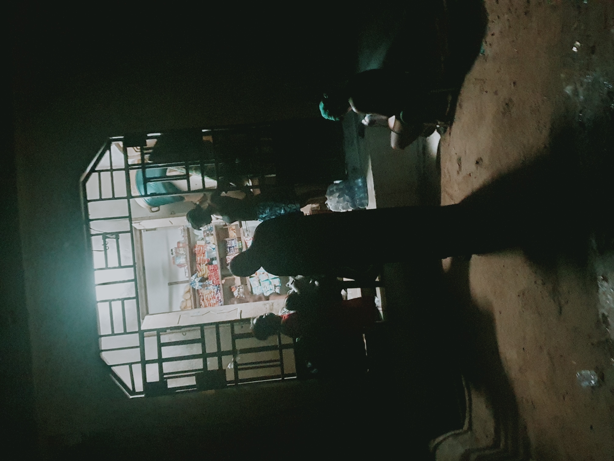 The image depicts an open-front shop in Lokoja, Nigeria, with several individuals present. A metal security grate frames the shop's opening. Inside the shop, shelves are stocked with assorted goods, including packaged snacks and beverages. Outside the shop, at least three people are visible; one appears to be making a purchase, while others are standing nearby. The scene is dimly lit, suggesting it is nighttime. The ground is dirt or unpaved. A water bottle is on a counter inside the shop.