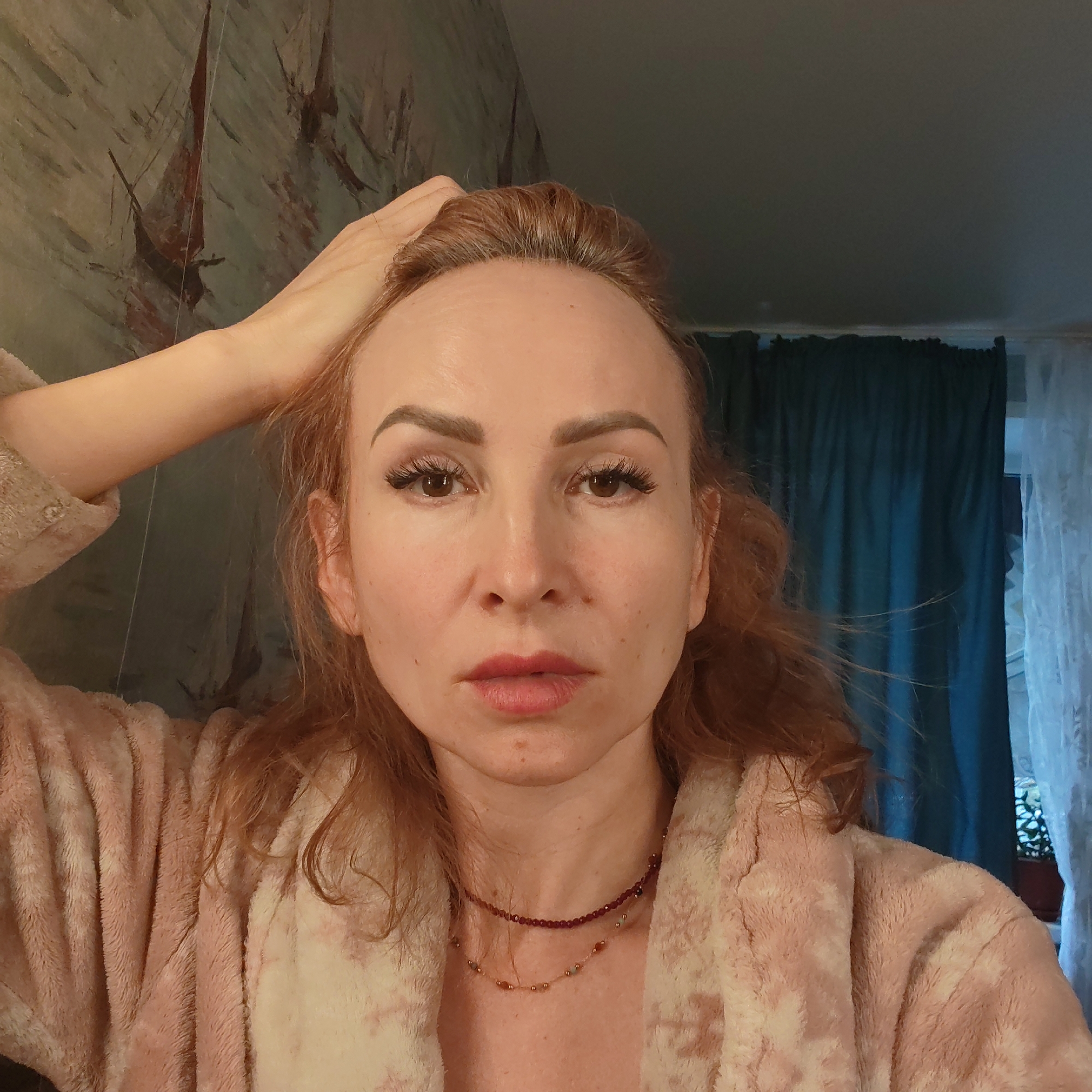 A woman with reddish-brown hair and fair skin stands in what appears to be an indoor setting. She is wearing a pink, plush bathrobe and a layered necklace. Her left hand is raised and touches her hair near her forehead. Her facial expression appears neutral.
Behind her, the wall is decorated with a mural featuring depictions of ships. A curtain is visible in the background near a window.
While the prompt suggests a location in Chaikovsky, Russia, this cannot be determined from the visual data.