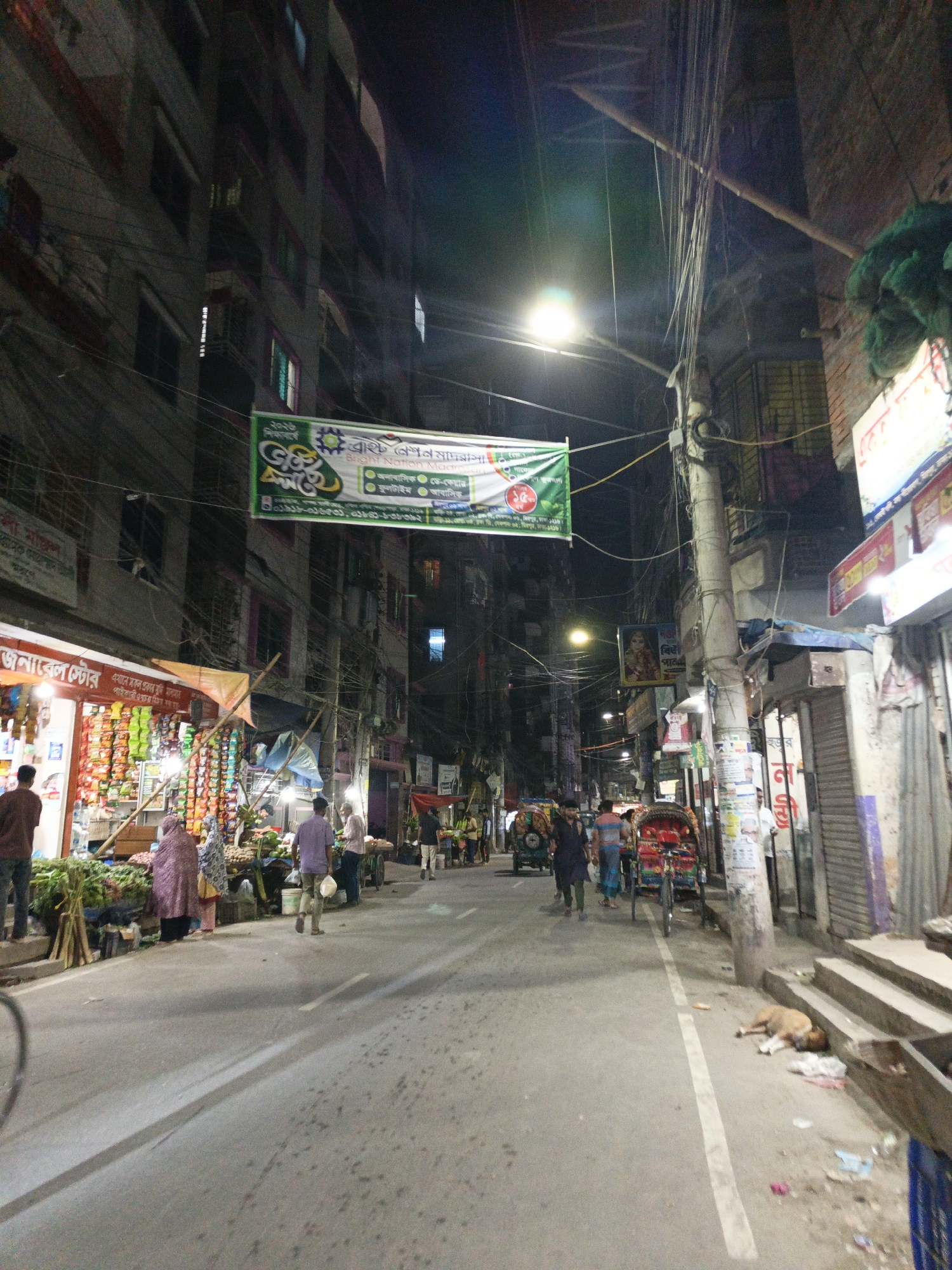 The image shows a street scene in Dhaka, Bangladesh, at night. Overhead are tangled electrical wires and streetlights illuminating the street. A banner hangs over the street with text in Bengali. On the left side of the street, there's a shop with goods displayed outside. People are walking along the street, and some are gathered near the shop. A few rickshaws are also visible. On the right side of the street, a dog is lying on the steps of a building. The buildings on either side of the street are multi-story and appear to be residential or commercial.
