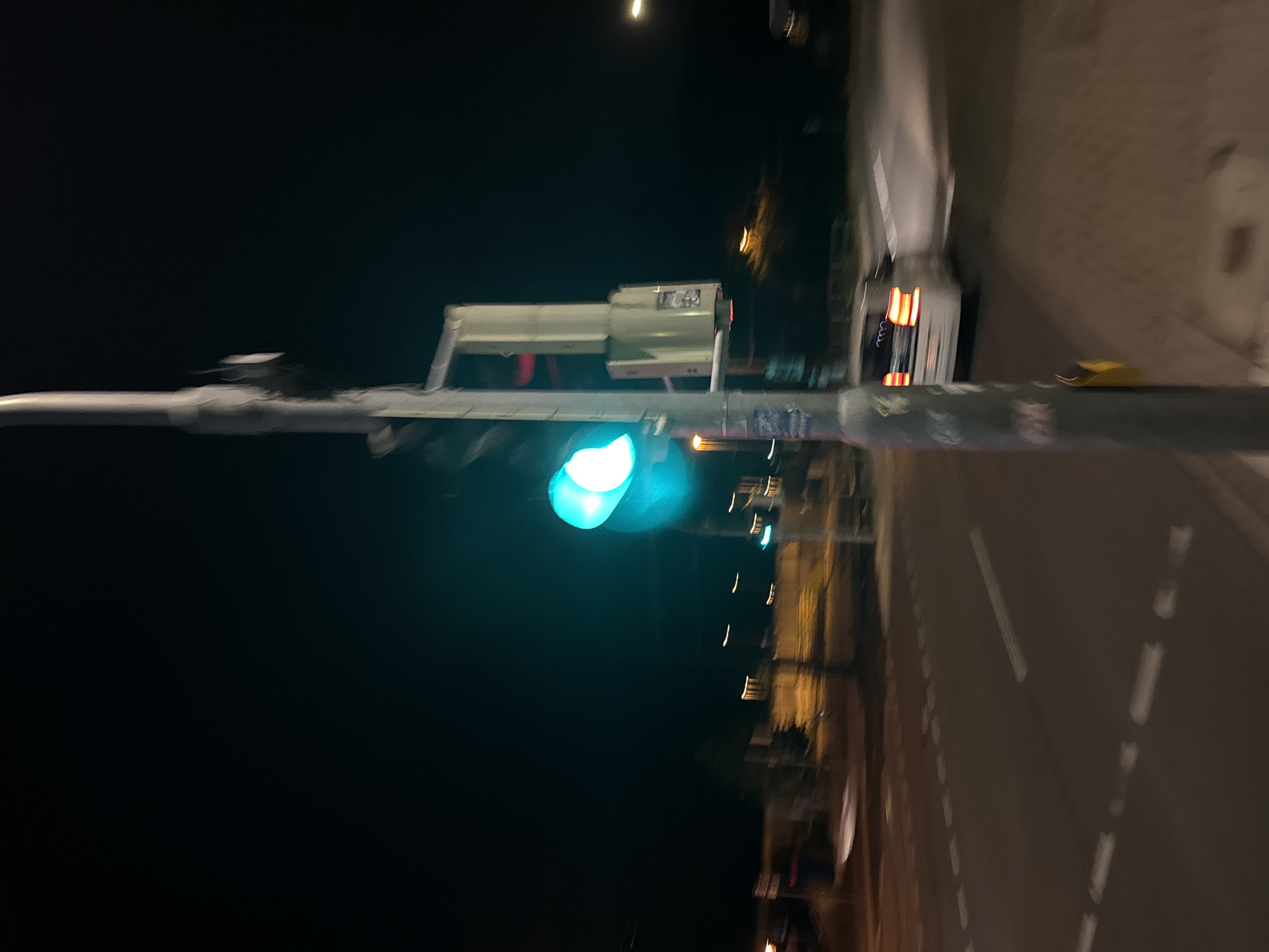 The image depicts a street scene at night in an unknown city. A traffic light is visible, illuminated with a green light. A white car with its taillights illuminated is turning right in the foreground. The scene appears to be an intersection with road markings visible on the asphalt. The overall image is slightly blurred, suggesting motion or low light conditions. There are no people clearly visible in the image. The environment appears to be an urban or suburban setting with streetlights and buildings faintly visible in the background.