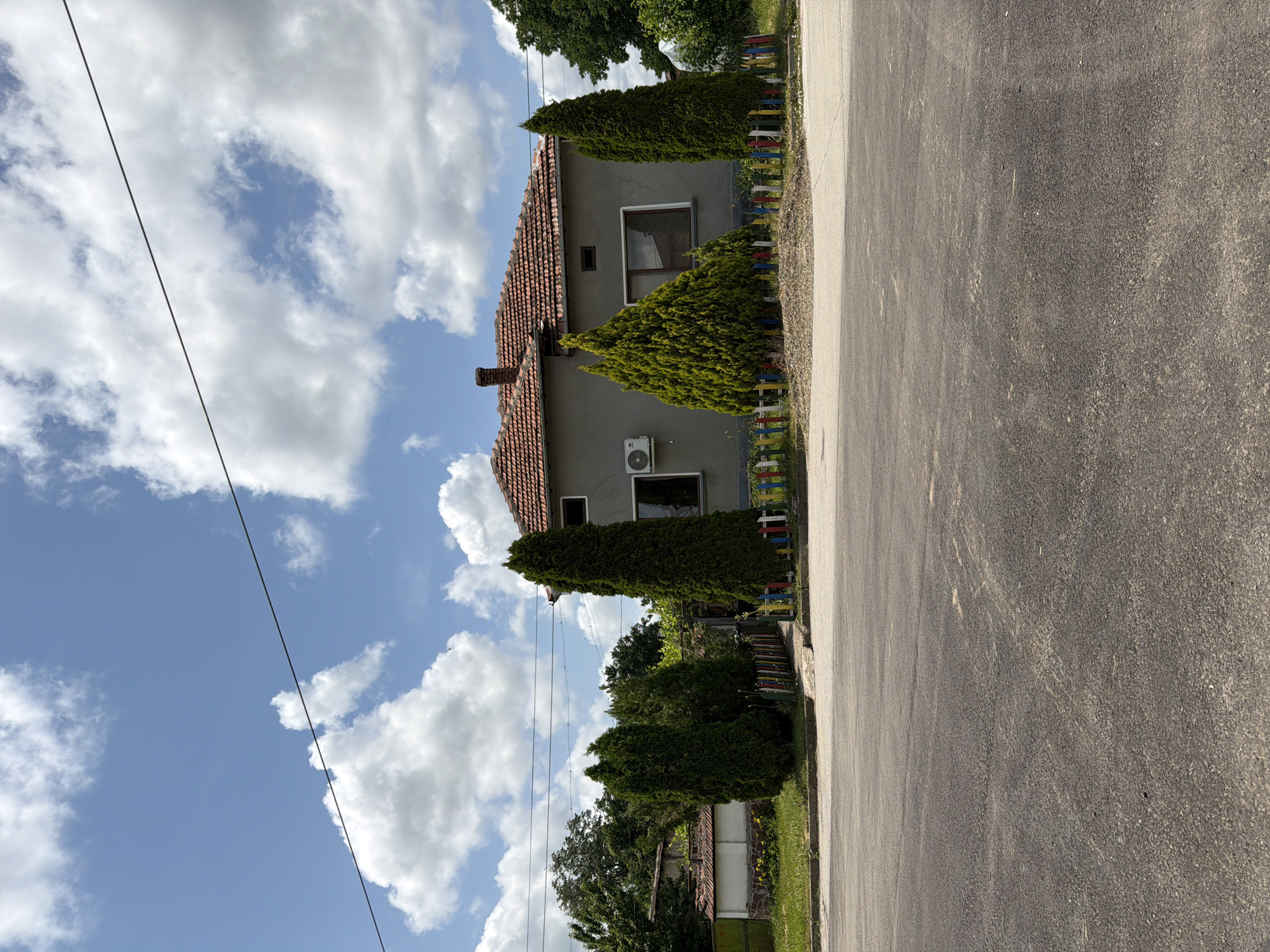 The image captures a residential scene from a slightly elevated perspective, looking across a paved road towards a house and its surrounding garden. The main subject is a one-and-a-half-story house with light-colored, possibly gray or beige, walls and a reddish-brown tiled roof. A chimney is visible on the left side of the roof, and an outdoor air conditioning unit is mounted below an upper-floor window. Another window is visible on the lower floor.
In front of the house, several tall, conical evergreen trees are neatly planted, bordering a low, brightly painted picket fence featuring multiple colors. Beyond the fence and house, more green trees and foliage are visible, indicating a lush, possibly rural or suburban environment.
The foreground is dominated by a dark gray paved road or street, showing some textural variations. Overhead, a bright blue sky is filled with numerous fluffy white cumulus clouds. Several black utility lines stretch diagonally across the upper left portion of the sky, passing above the house.
No people are visible, and no specific activity is taking place, presenting a tranquil, static scene. The clear sky and bright lighting suggest a sunny day, likely mid-morning or afternoon. The overall setting, including the house architecture and vegetation, is consistent with a village or small town environment, aligning with the provided location context of Dermantsi, Bulgaria. There is no visible text in the image.