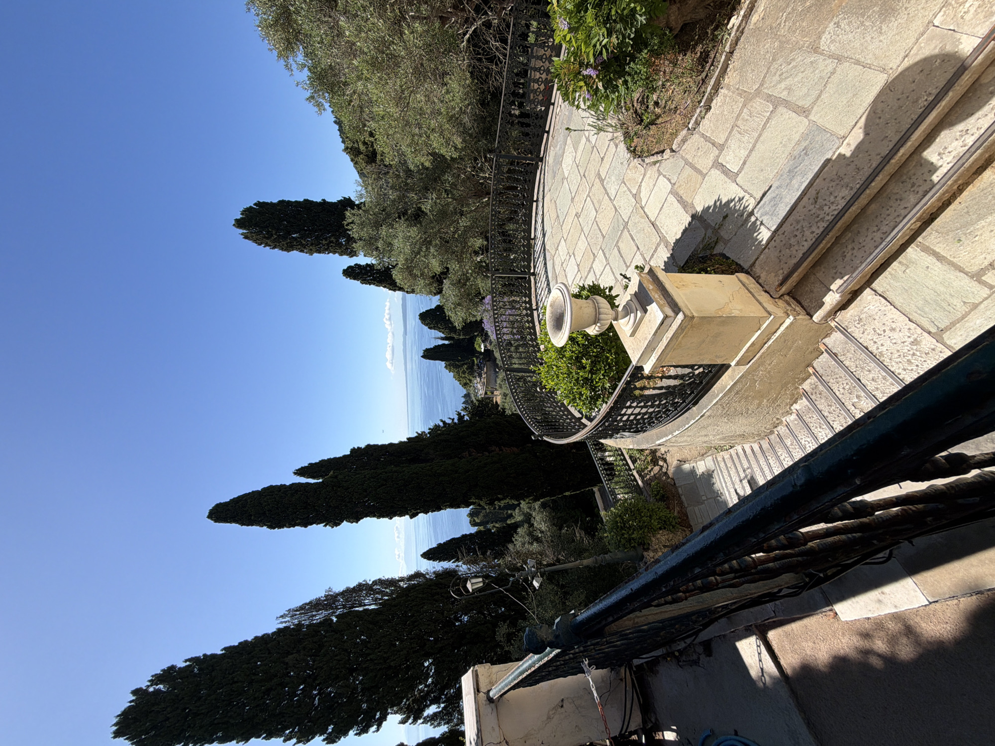 The image captures a picturesque outdoor garden or terraced area with a panoramic view of the sea under a brilliant clear sky. The scene is oriented as if looking down and across a multi-level landscape.
In the foreground, a light-colored, patterned stone path or patio leads to stone steps on the right. A dark, ornate wrought-iron railing runs along the edge of these steps and a low wall, contributing to the classical aesthetic. A prominent stone pedestal with a classical urn sits on the right, beside the path. Shadows from the railing and nearby foliage are cast sharply onto the stone, indicating strong sunlight.
Extending into the midground, a gracefully curved stone path, also paved with patterned stones, follows the contours of the land. This path is bordered by a continuous, dark wrought-iron railing, offering a vantage point over a drop-off or lower garden section. Lush green vegetation abounds, featuring several tall, slender, dark green cypress trees that frame the vista and other bushy greenery lining the paths and garden beds.
The background reveals a vast, tranquil expanse of deep blue sea stretching horizontally to meet a clear, bright blue sky. A faint, distant landmass or coastline is visible across the water on the horizon. The sky is almost entirely cloudless, reinforcing the impression of a beautiful, sunny day.
The scene is devoid of people, evoking a sense of peaceful solitude and natural beauty. The combination of cypress trees, stone architecture, and the Mediterranean sea view strongly suggests a location in a warm, coastal climate, consistent with Gastouri, Greece. The lighting implies it is either morning or late afternoon. No text is visible in the image.