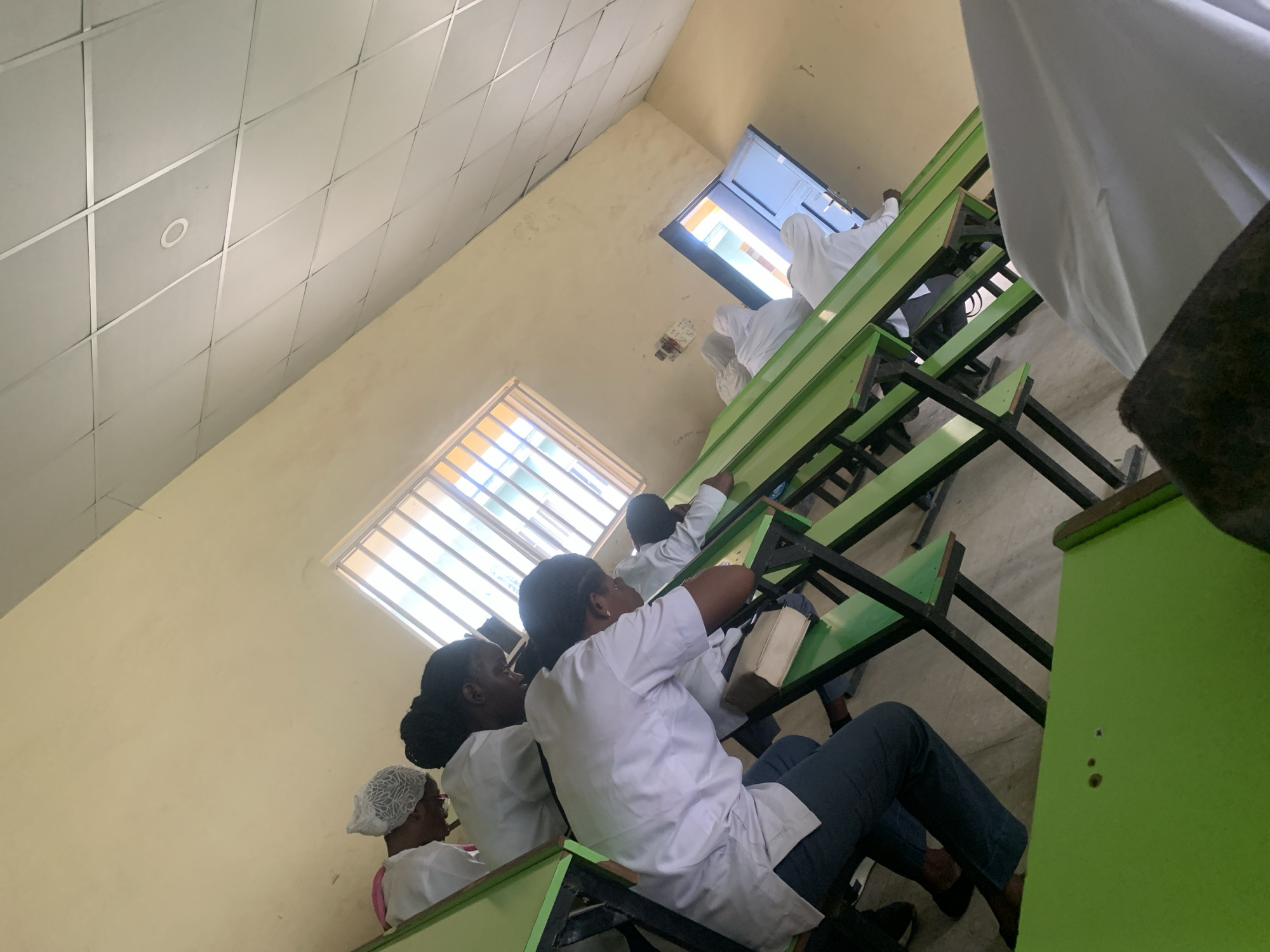 The image shows an indoor classroom setting in Keffi, Nigeria. Several individuals are seated at green and black desks. They appear to be wearing white lab coats, suggesting they might be students in a medical or science-related field. There's a window with metal bars and another open one. The walls are painted in a light yellow color. The room is lit by natural light coming through the windows and a ceiling light. The view is taken at a slight angle from one side of the room.