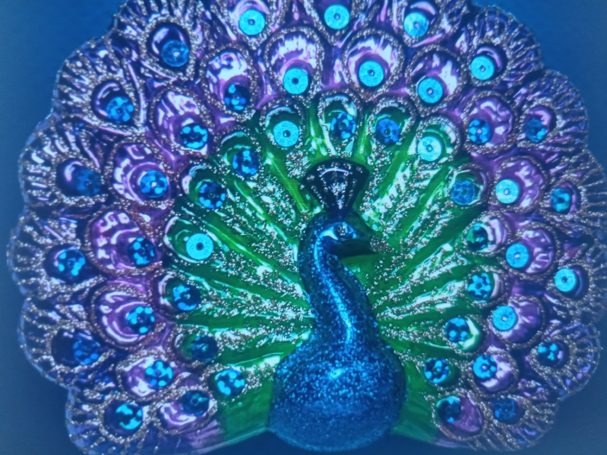 This is a close-up, slightly blurry, top-down shot of a decorative peacock ornament. The peacock's body is a vibrant, glittery blue. Its neck is elegantly curved upwards, and its head is pointed slightly to the left. The peacock's tail feathers fan out widely, forming a semicircle behind it. The feathers are detailed and layered, with a shimmering gradient of green and purple. Each feather is adorned with a distinct "eye" pattern, featuring a circular, bright blue gem or sequin at its center, surrounded by intricate metallic gold and purple embellishments. The overall impression is one of dazzling, artificial beauty, likely intended for display or decoration. The background is a plain, dark blue, which helps the colorful peacock stand out. There is no visible text or any indication of time of day, weather, or specific location. The image focuses entirely on the ornamental object itself.