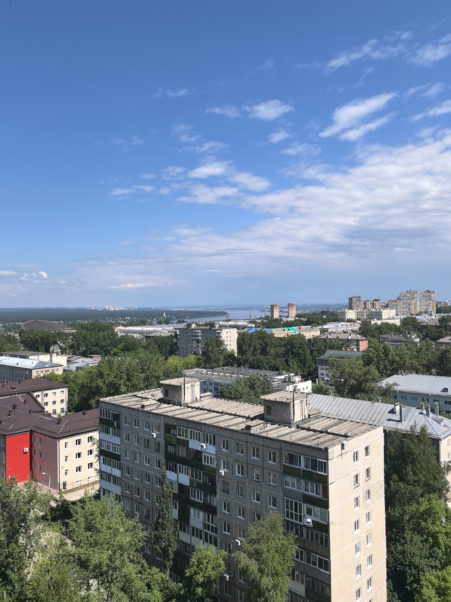 This is an elevated view of a cityscape in Perm, Russia. The sky is blue with scattered white clouds. Below, a dense cluster of trees surrounds a mix of residential buildings. One building stands out with a red facade, contrasting with the predominantly grey concrete structures. In the distance, there's a body of water and more buildings on the horizon, indicating the urban sprawl of Perm. The overall impression is of a city embedded in lush greenery.