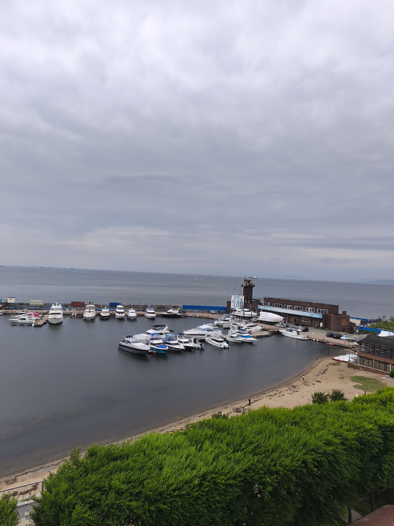 The image shows a marina with numerous yachts and smaller boats docked in the foreground, overlooking a calm body of water. In the background, several larger ships are visible on the horizon. A brick building with a tall tower, likely a lighthouse or observation post, stands to the right of the marina. A sandy beach is situated between the marina and a dense line of green trees that occupy the bottom of the frame. The sky is overcast, suggesting a cloudy day. The overall scene depicts a coastal area within the city of Vladivostok, Russia, characterized by its maritime activities and urban development alongside natural elements.