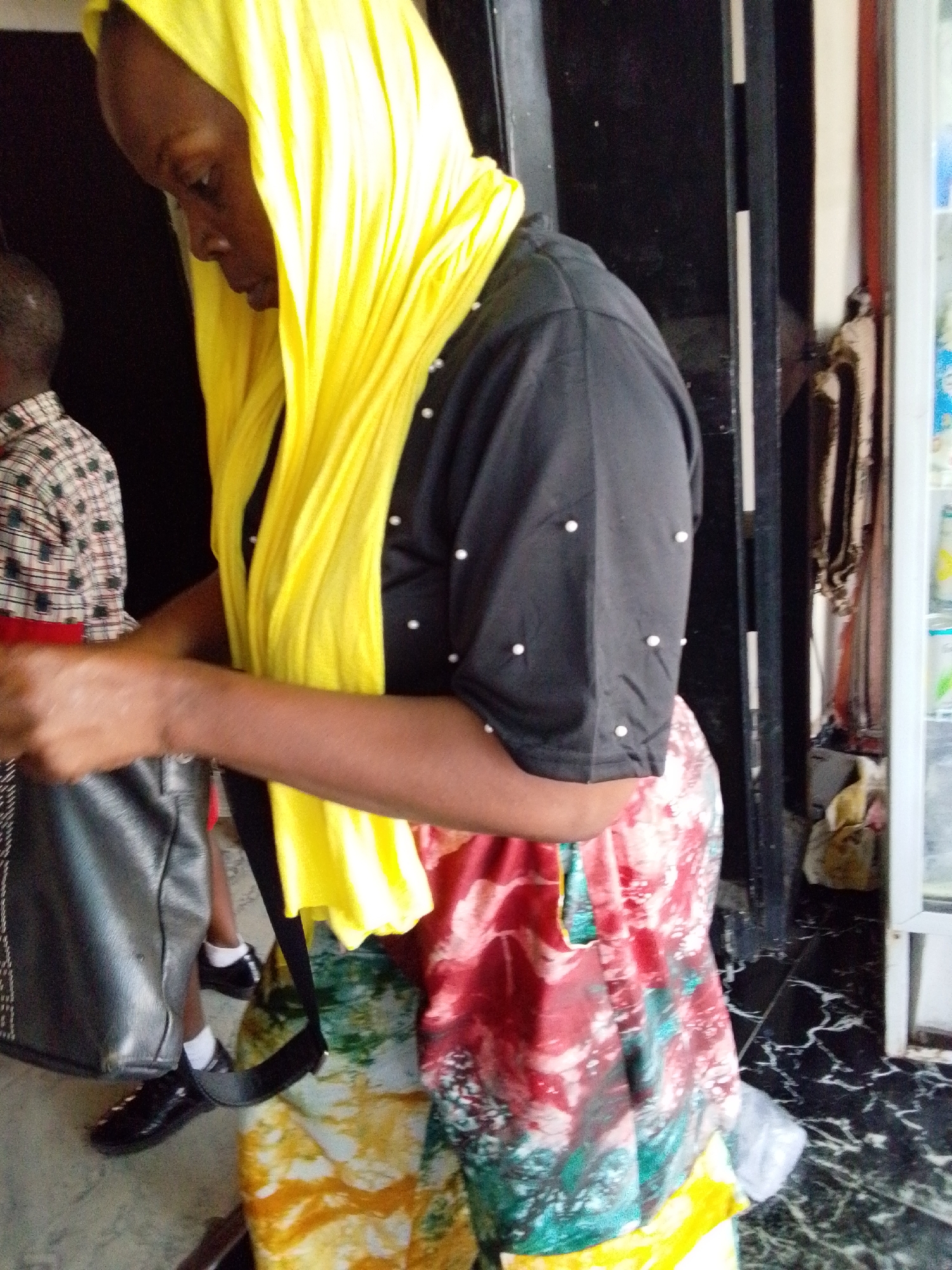 A woman with dark skin and short hair is wearing a bright yellow headscarf, a black blouse with small white embellishments, and a colorful red, green, yellow, and white patterned skirt. She is standing near a doorway, possibly a shop entrance in Lokoja, Nigeria. Next to her is a person in a plaid shirt and dark pants, carrying a large dark-colored bag. The floor has a black and white marble pattern. Behind her is a black structure with white trim, and a glass-fronted display case. The overall lighting is dim, suggesting an indoor setting.