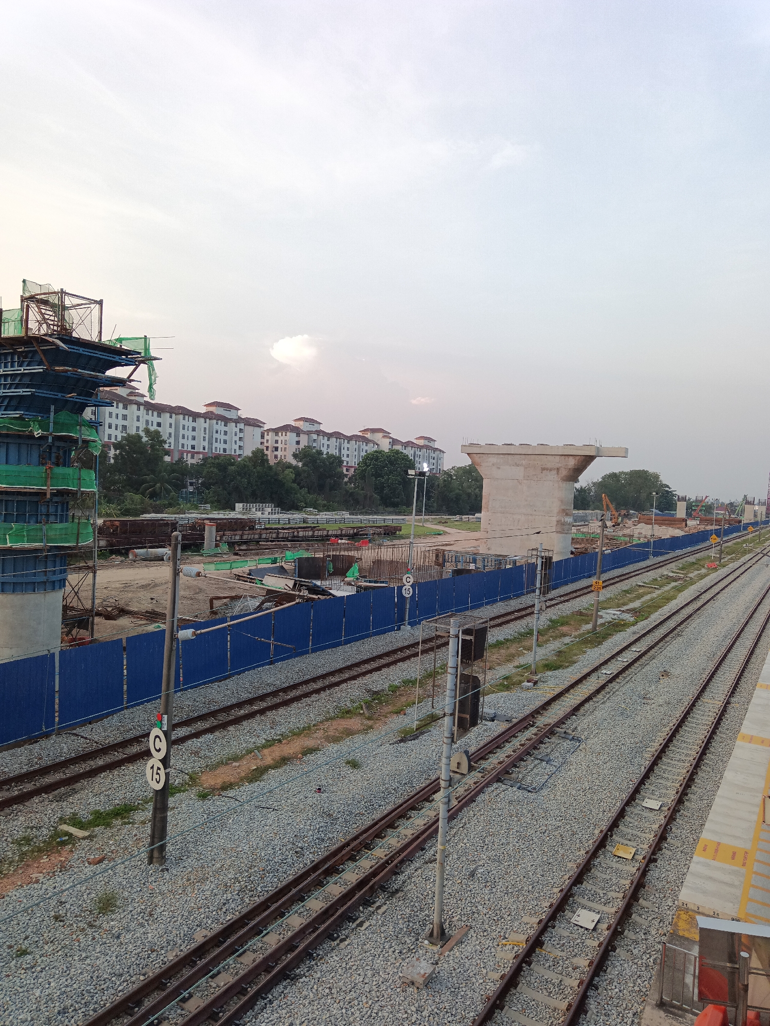 This is a wide-angle, eye-level outdoor photograph of a construction site adjacent to railway tracks in Klang City, Malaysia. The sky is overcast with a pale, hazy light, suggesting it is either early morning or late afternoon.
In the foreground, multiple sets of parallel railway tracks stretch into the distance, covered with gravel. A concrete platform with yellow markings is visible on the far right. A utility pole with signals and a circular sign with the letter "C" and the number "15" is positioned near the tracks. A blue construction fence runs along the left side of the railway lines, separating them from the construction area.
The construction site itself is extensive and features various elements. On the far left, a large, multi-story structure, possibly part of an elevated railway or bridge, is under construction. It is made of blue scaffolding, concrete pillars, and green netting. Further into the site, a prominent concrete structure resembling a bridge support column stands. In the background, several rows of residential buildings with light-colored walls and reddish-brown roofs are visible, separated by a line of trees. Various construction materials, machinery, and smaller structures are scattered throughout the site, including what appear to be concrete beams and excavated earth. The overall impression is one of ongoing infrastructure development in an urban or semi-urban setting.