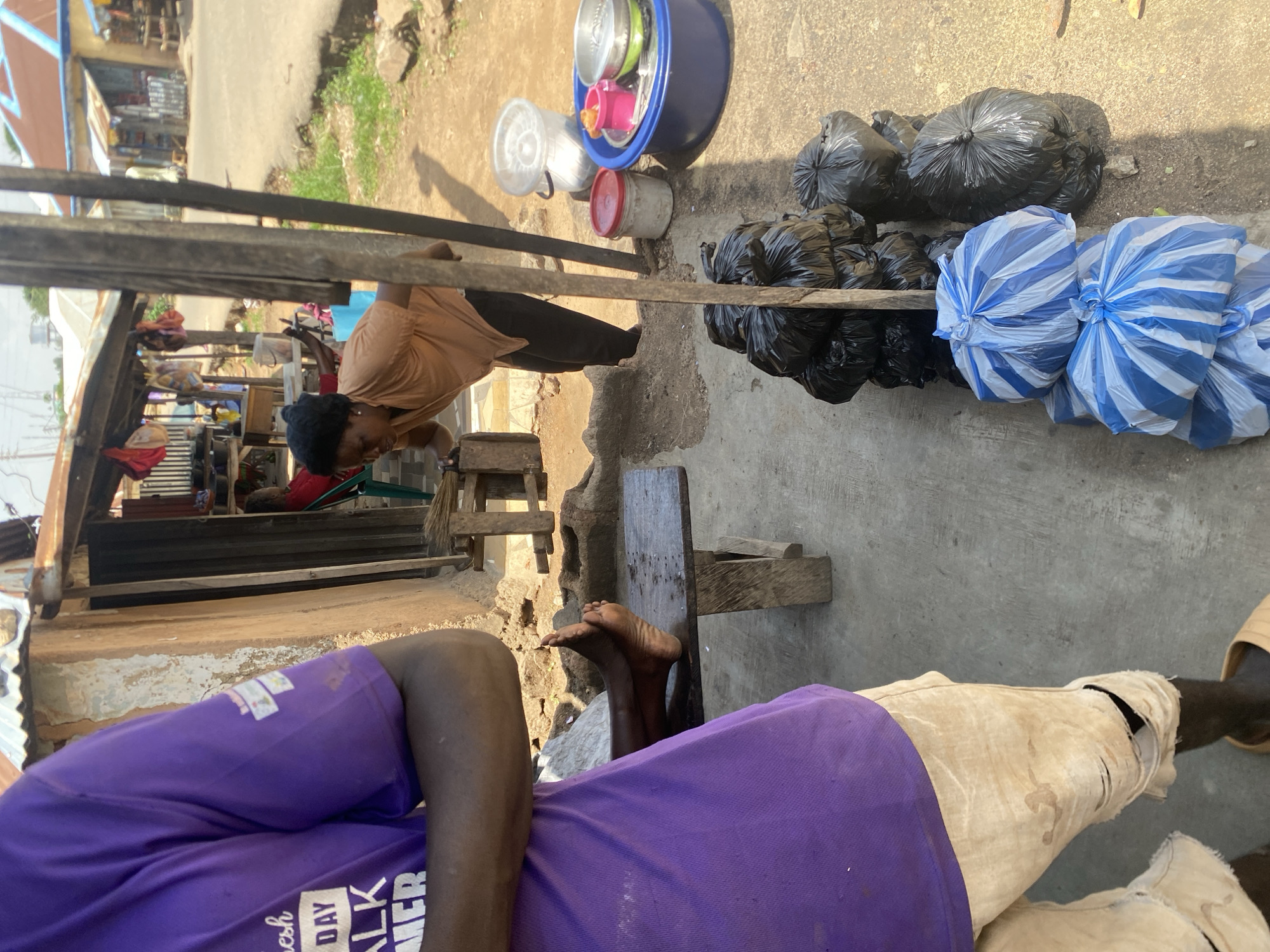 The image depicts an outdoor scene in Jos, Nigeria, likely in a market or roadside stall area. In the foreground, a person wearing a purple t-shirt and tan shorts with rips is partially visible, with their feet resting on a wooden bench. To the right, a woman in an orange shirt is leaning over, appearing to interact with goods or a structure. Behind her, several black trash bags are stacked, along with some blue and white striped bags. Various containers, including buckets and bowls, are also present, some appearing to hold items. The background shows simple structures with corrugated metal roofs, a dirt path, and a paved road with some vegetation. The overall impression is one of a bustling, informal commercial environment.