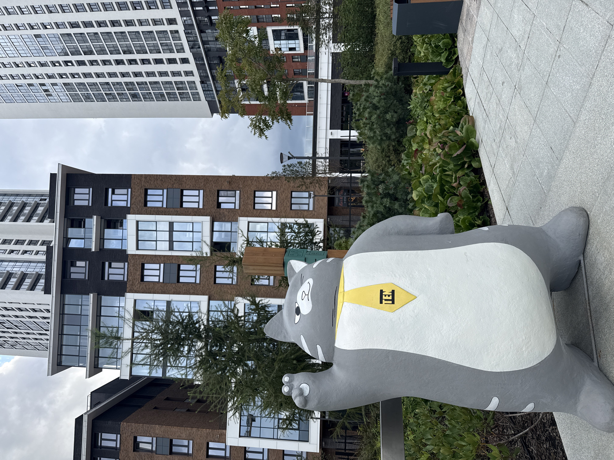 A gray, cartoonish cat sculpture wearing a yellow tie with the "TEN" logo stands prominently in the foreground of the image. The sculpture is positioned on a paved walkway, with lush green bushes and trees surrounding it. In the background, modern apartment buildings with a mix of brick and glass facades rise against a cloudy sky. The architecture suggests a contemporary urban development. The scene is located in the residential complex "Forest Park" in Yekaterinburg, Russia.