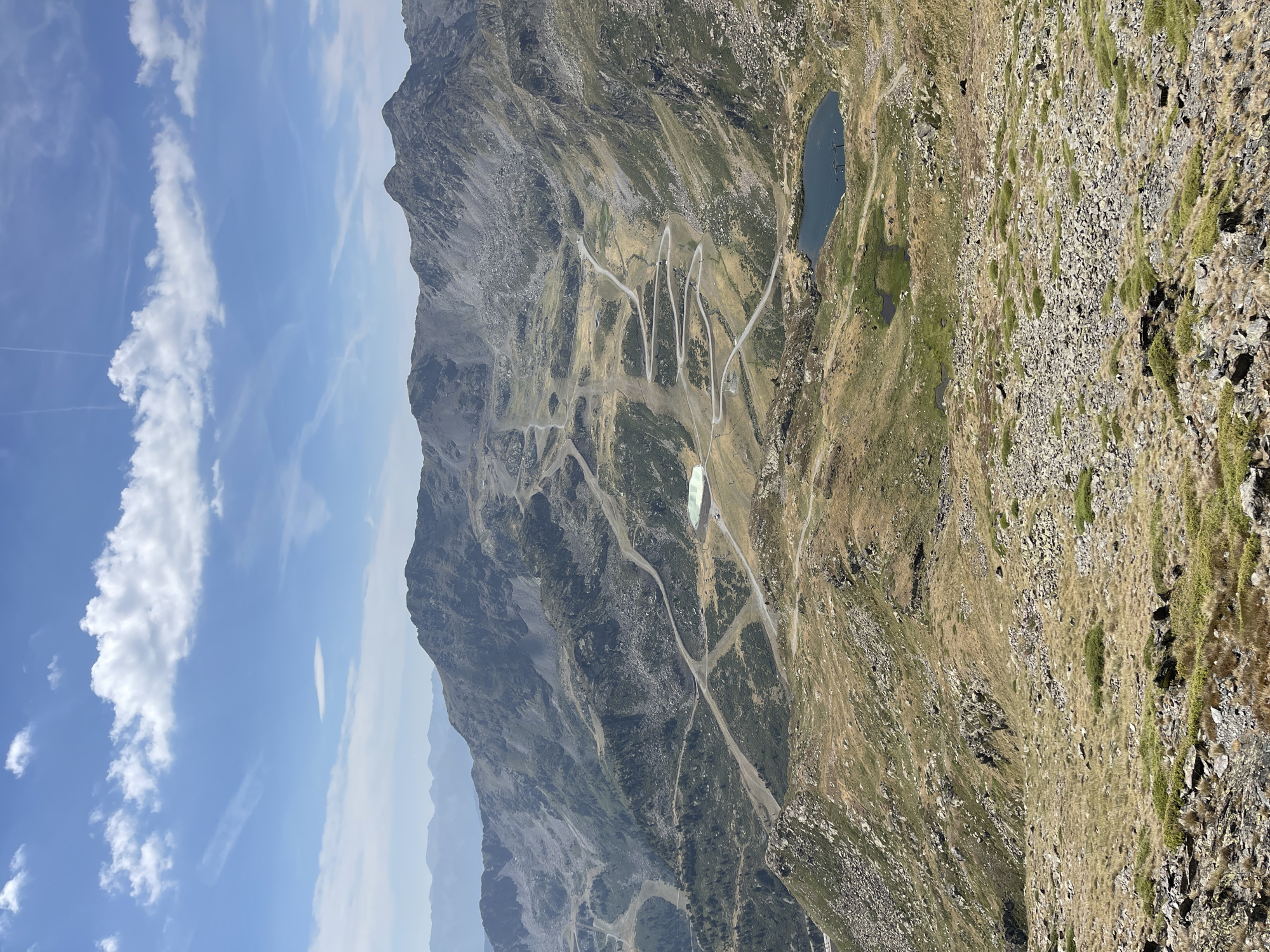 The image captures a sweeping, high-altitude mountain landscape under a bright, partly cloudy sky. The terrain is rugged and mountainous, with steep slopes covered in a mix of rocks, dry grass, and scrubby vegetation. Numerous winding roads, some paved and some unpaved, snake their way up and across the mountainsides, indicating a developed mountain resort or pass. Several ski slopes are also visible, marked by cleared paths and possibly ski lifts.
In the middle ground, two distinct bodies of water are present. The first is a large, pale green, man-made reservoir or pond, likely used for snowmaking, with a small structure or building at its edge. Further down, a smaller, deep blue natural lake is visible, with what appears to be ski lift infrastructure on its shore. The distant mountains fade into a hazy blue, suggesting a vast and expansive panorama.
The foreground is dominated by a rocky, barren slope with sparse, dry grass and scattered larger stones. The overall impression is one of a remote, natural environment that has been significantly shaped by human activity for recreation and infrastructure. There are no people visible in the image. The scene is illuminated by bright sunlight, casting sharp shadows on the rugged terrain.
